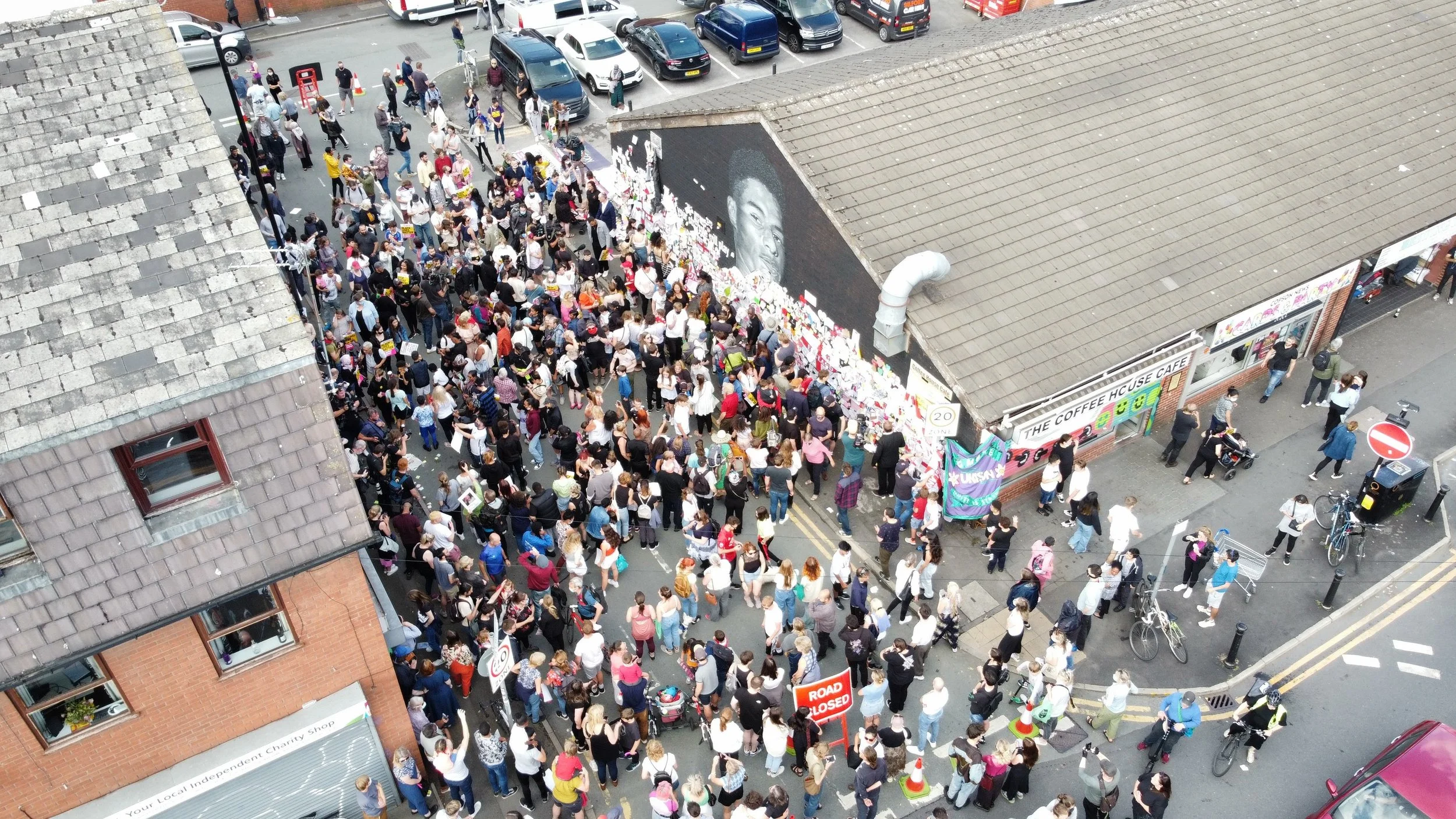 Drone view of a large crowd infront of the Marcus Rashford mural in Withington. The mural is covered in plags, messages and other items paying tribute.