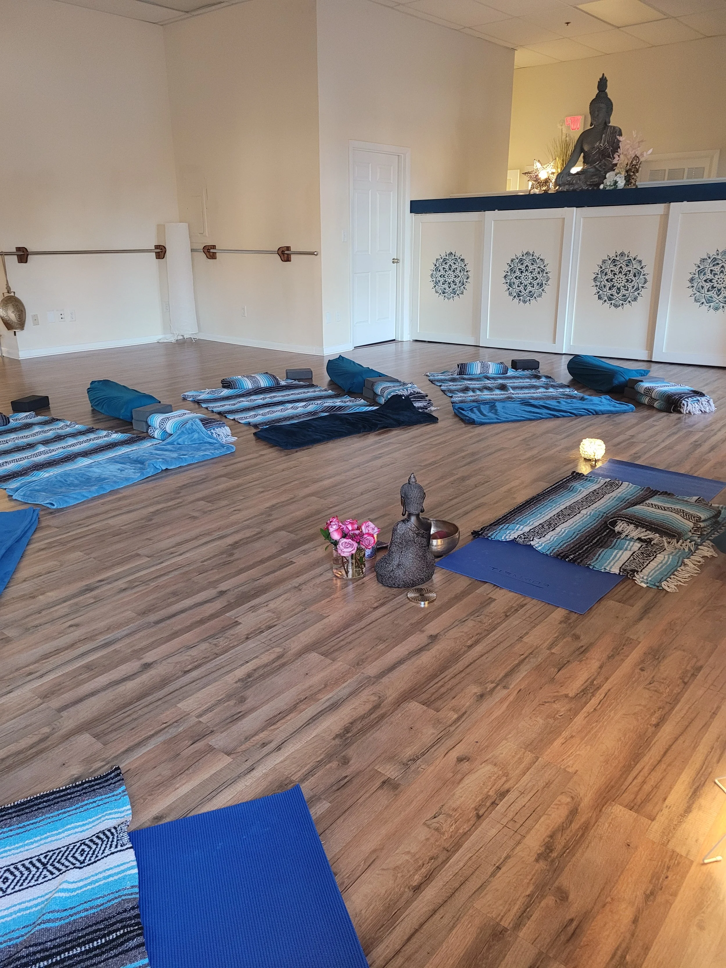Yoga setup with mats and pillows arranged on a wooden floor in a spacious room, decorated with a Buddha statue, flowers, and ambient lighting.