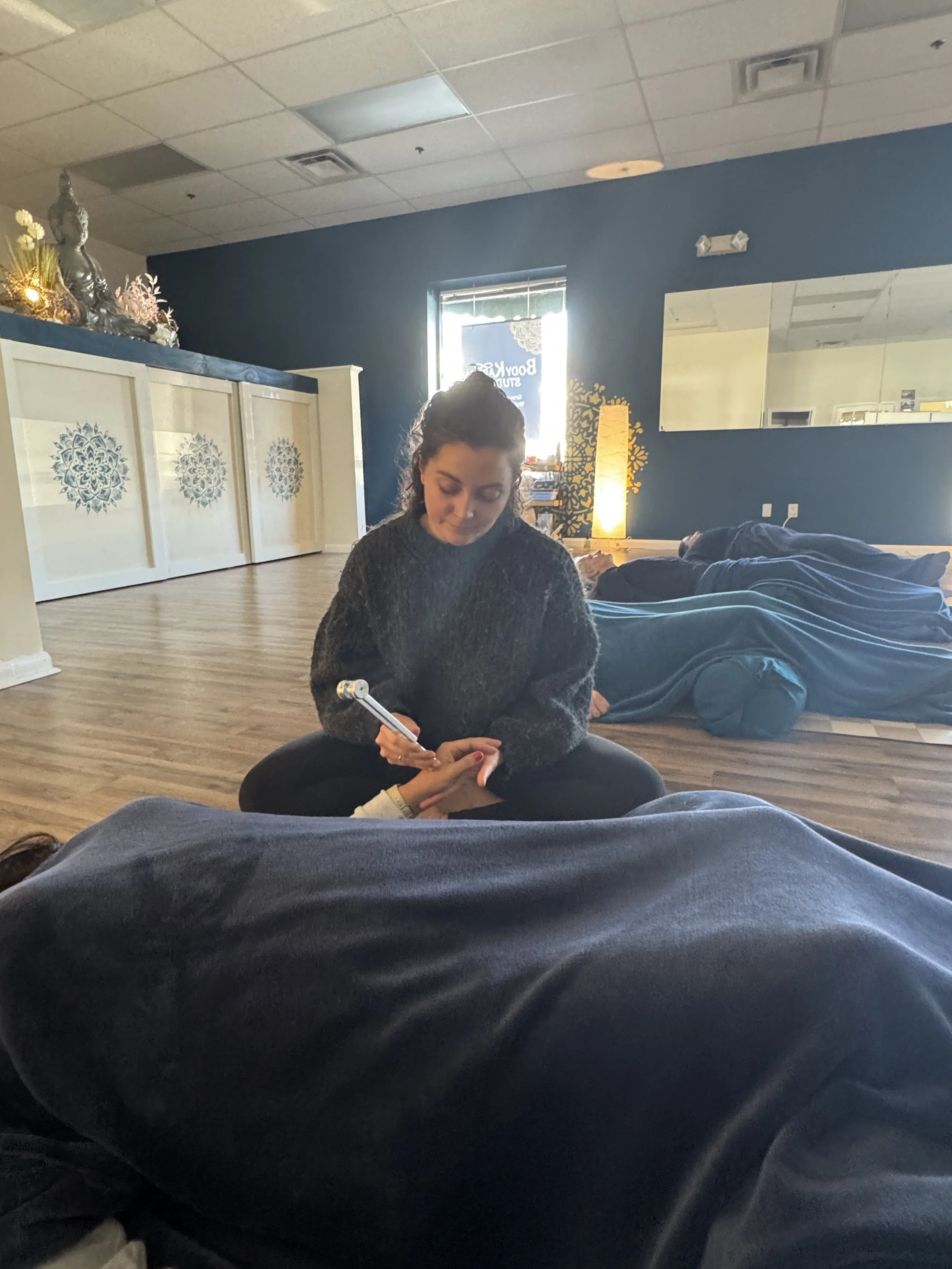 A woman sitting on the floor receiving a foot massage in a wellness studio with blue walls, wooden flooring, and decor including a Buddha statue on a ledge and a decorative tree silhouette mirror.