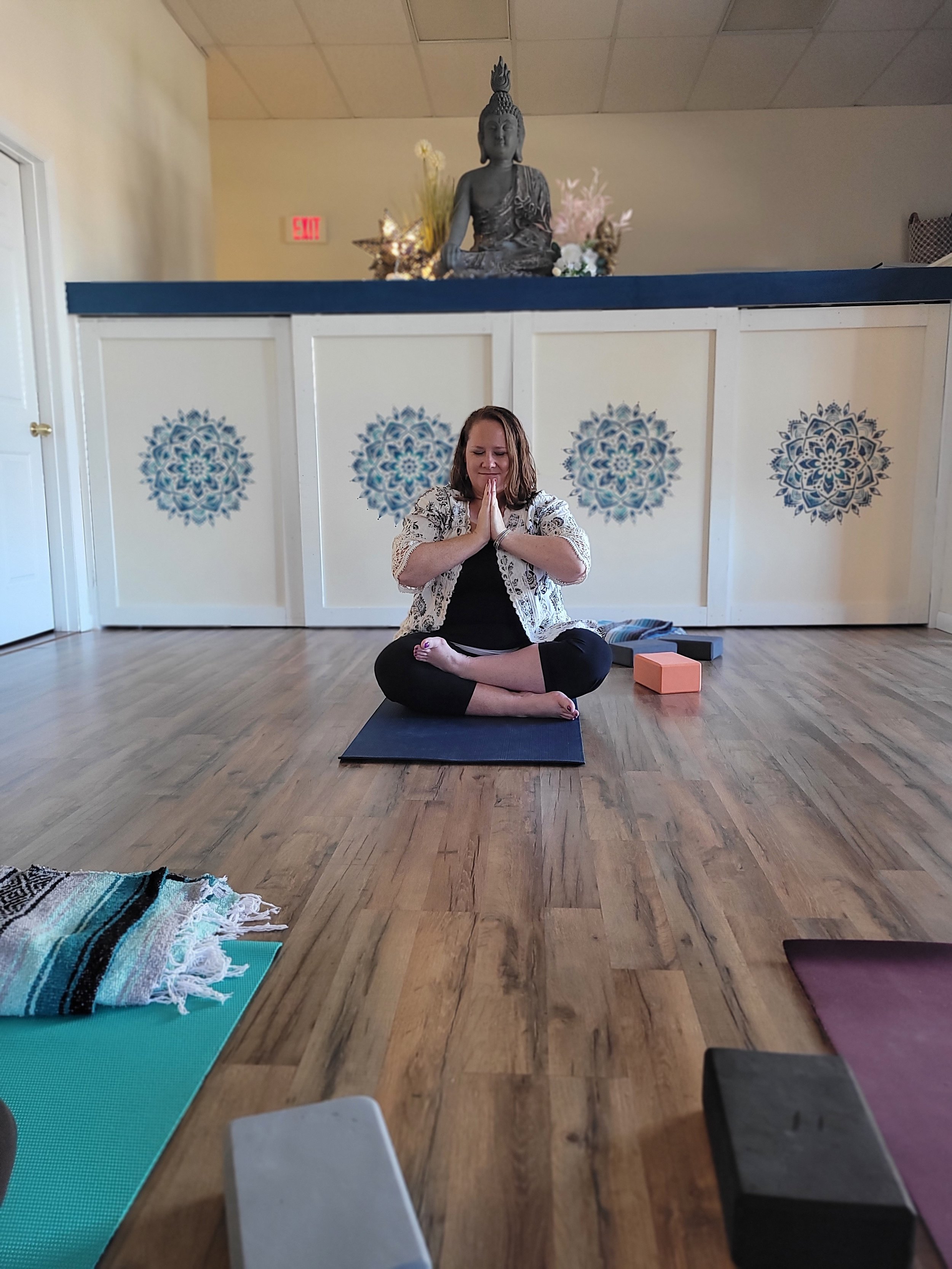 A woman practicing yoga or meditation in a room with a Buddha statue on a decorated altar. She is seated on a yoga mat with her eyes closed and hands in prayer position.