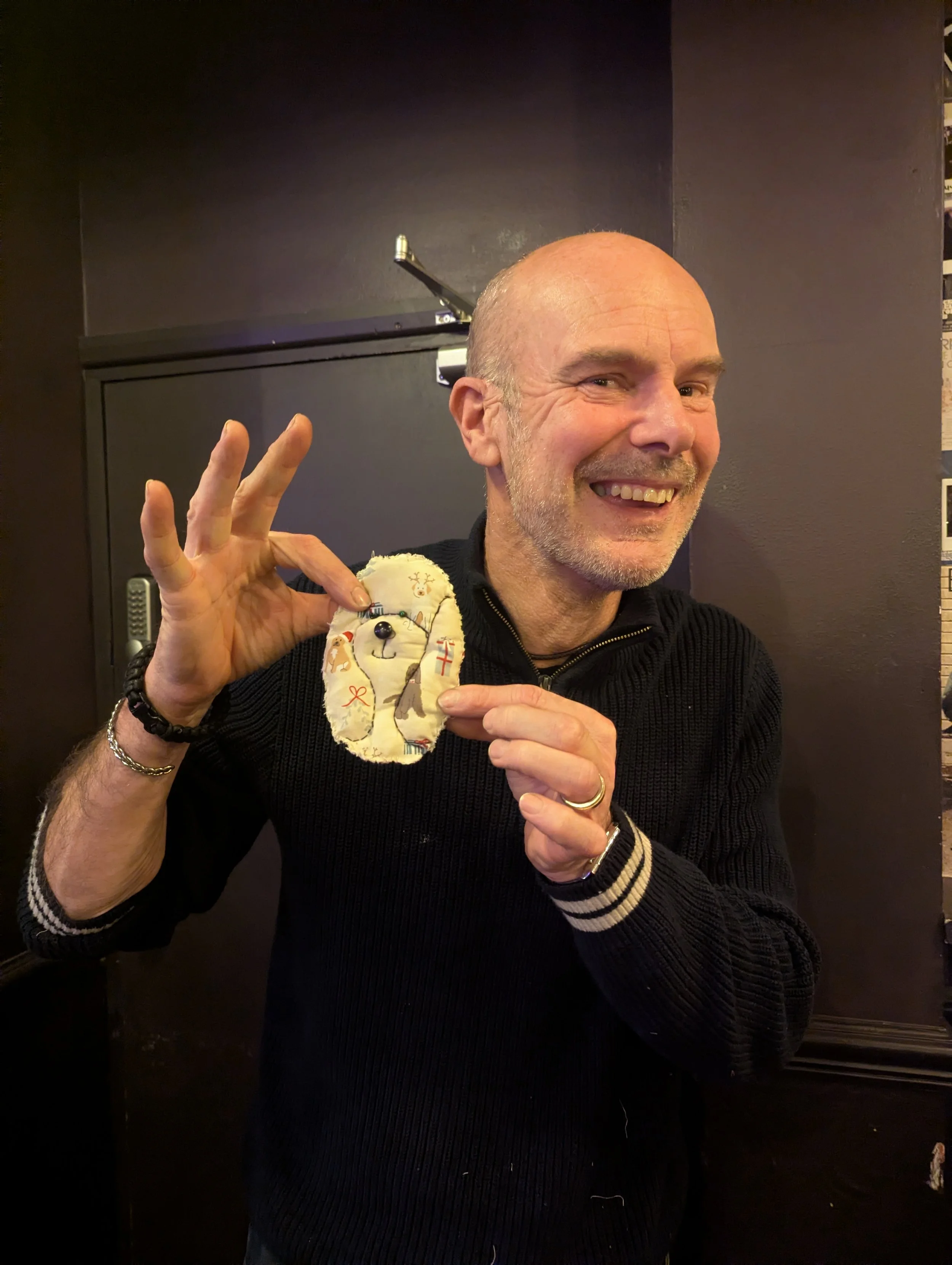 A smiling man holding a fabric dog plush toy and making an OK sign with his right hand. The man has a short beard and is wearing a black sweater with striped cuffs.