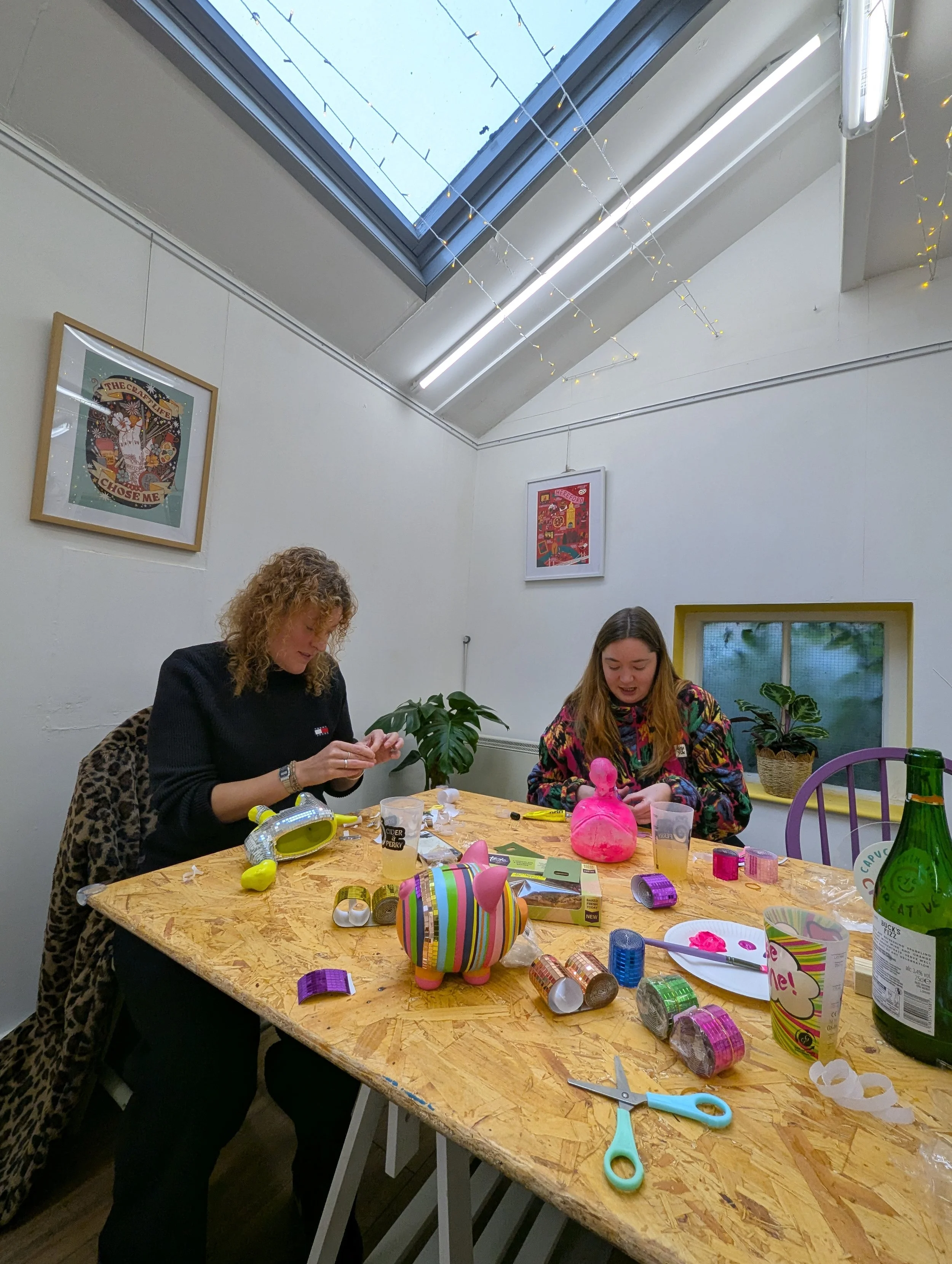 Two women sitting at a wooden table celebrating, surrounded by colorful ribbons, tape rolls, scissors, a pink balloon sculpture, and a large green bottle, with fairy lights and framed artwork on the white walls near a skylight.