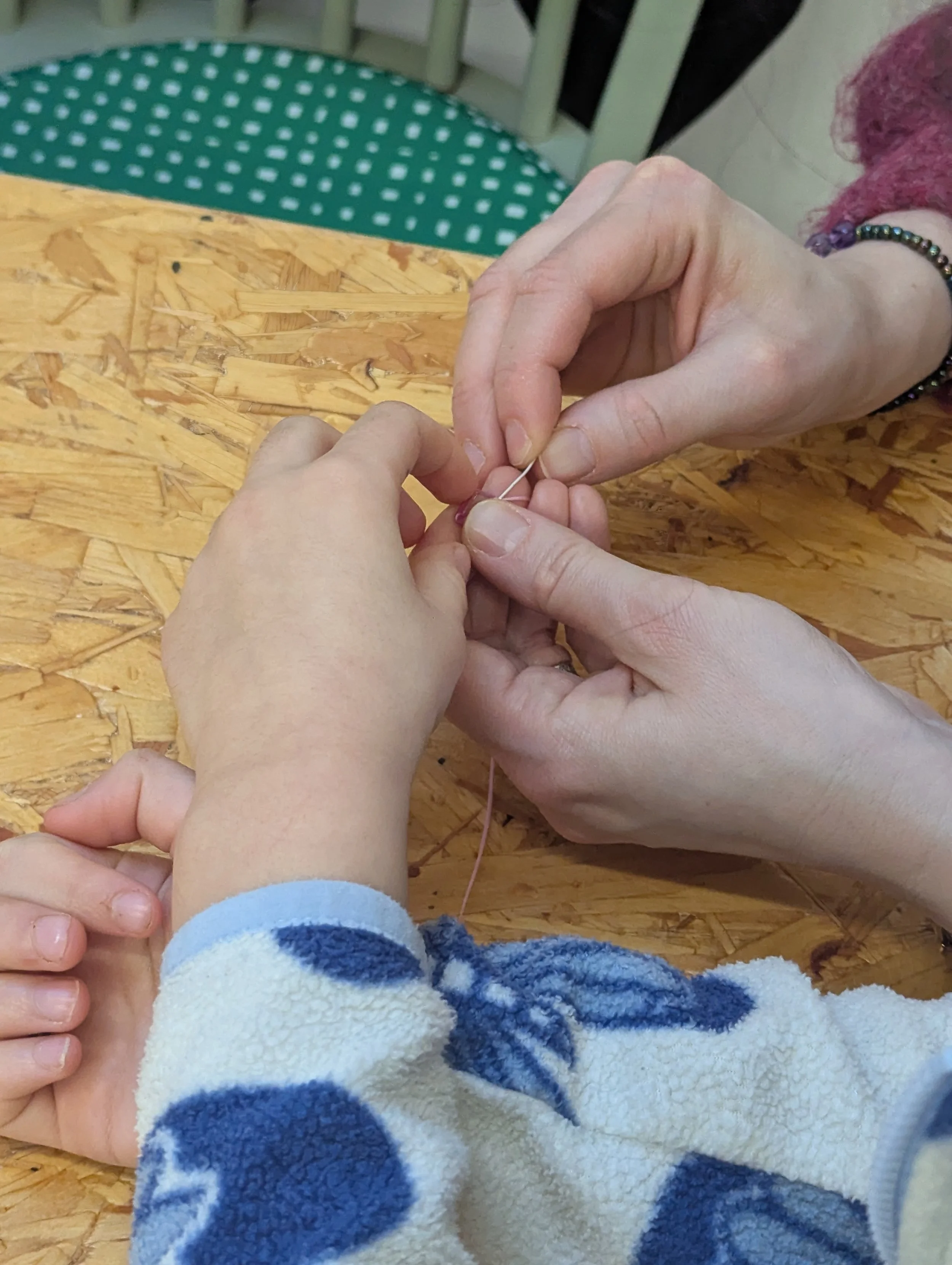 Two people sewing a small piece of fabric together using a needle and thread, with a child's hand visible.