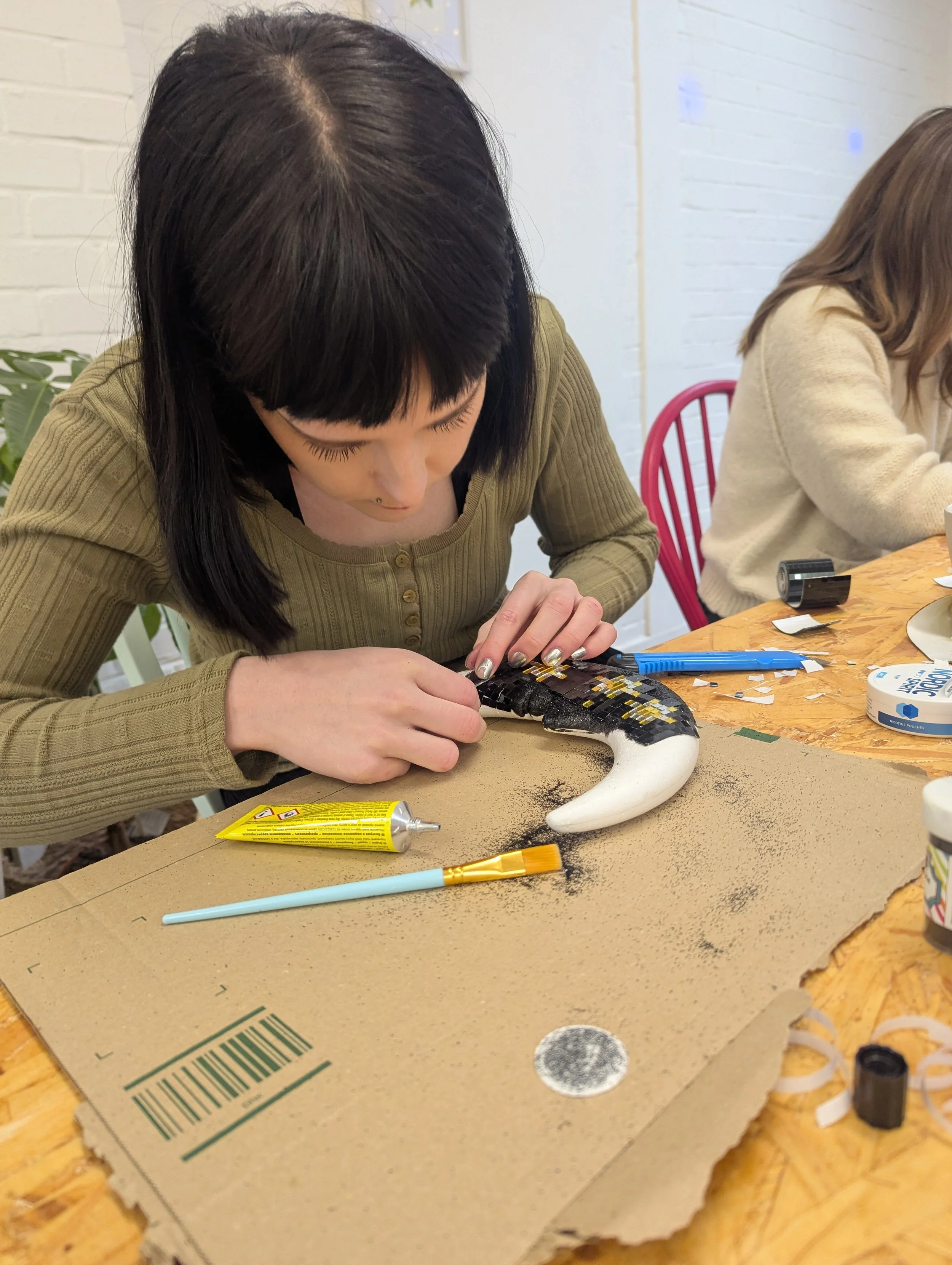 A woman with black hair and light skin is decorating a crescent moon-shaped object with black and yellow mosaic pieces at a table. There are craft supplies like glue, a brush, tape, and paper scattered on the table, with a woman in a beige sweater wo
