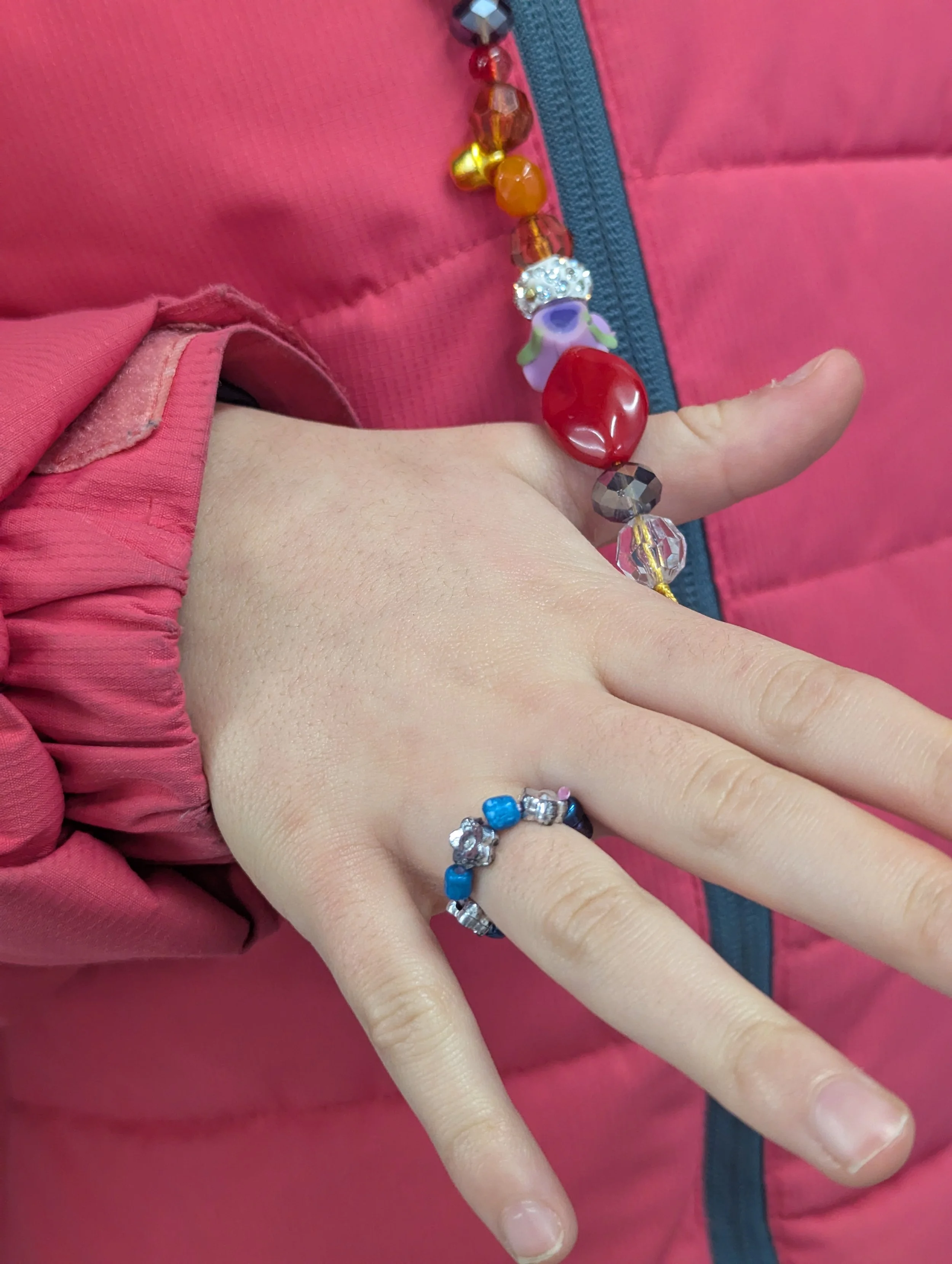 Close-up of a person's hand wearing a beaded bracelet and a ring, with the background showing a pink jacket and colorful beaded necklace.