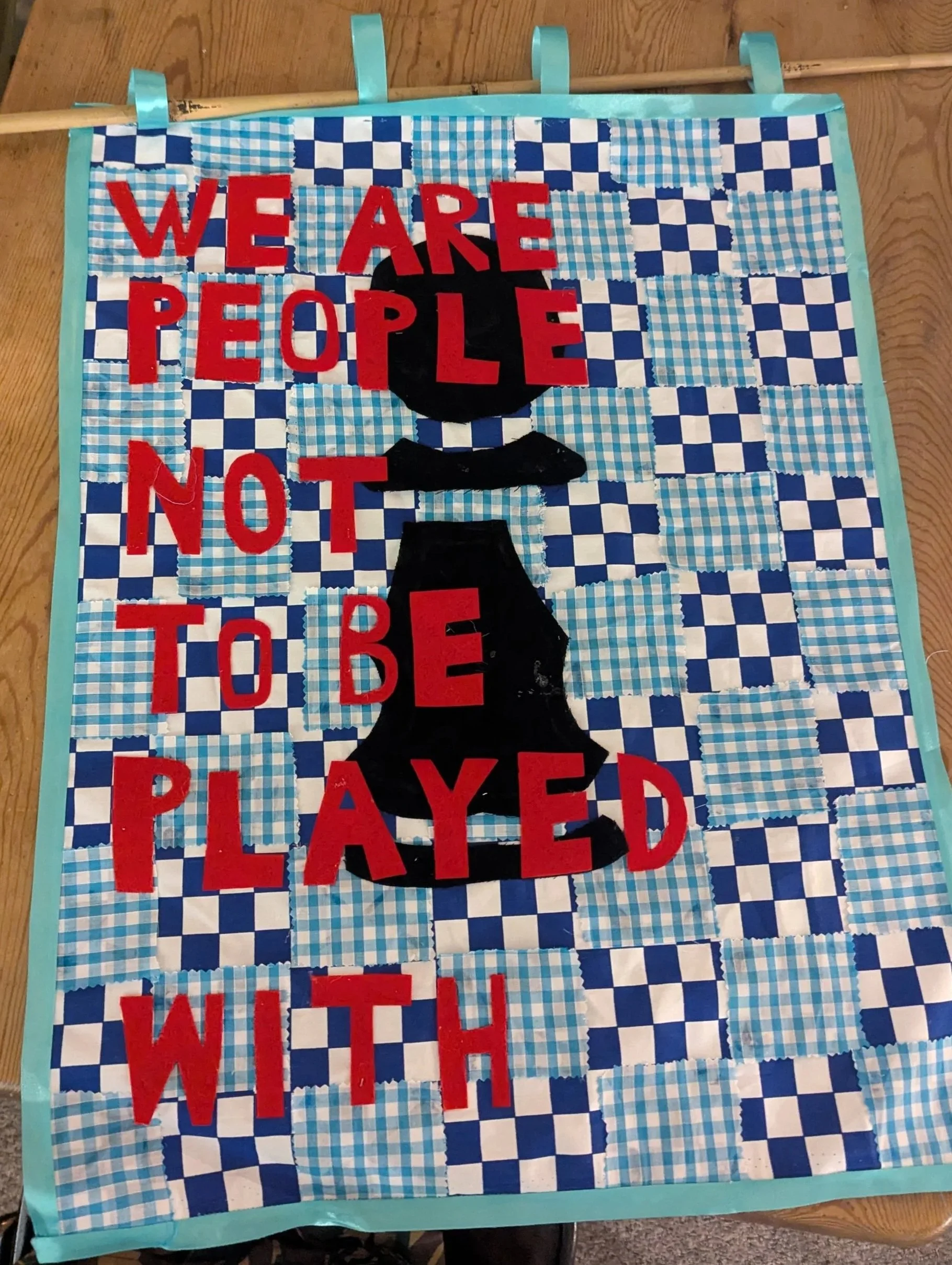 A quilt with a blue and white checkered pattern as background, featuring black cutouts of a trophy and a baseball cap, with red felt letters forming the phrase: 'WE ARE PEOPLE, NOT TO BE PLAYED WITH.'