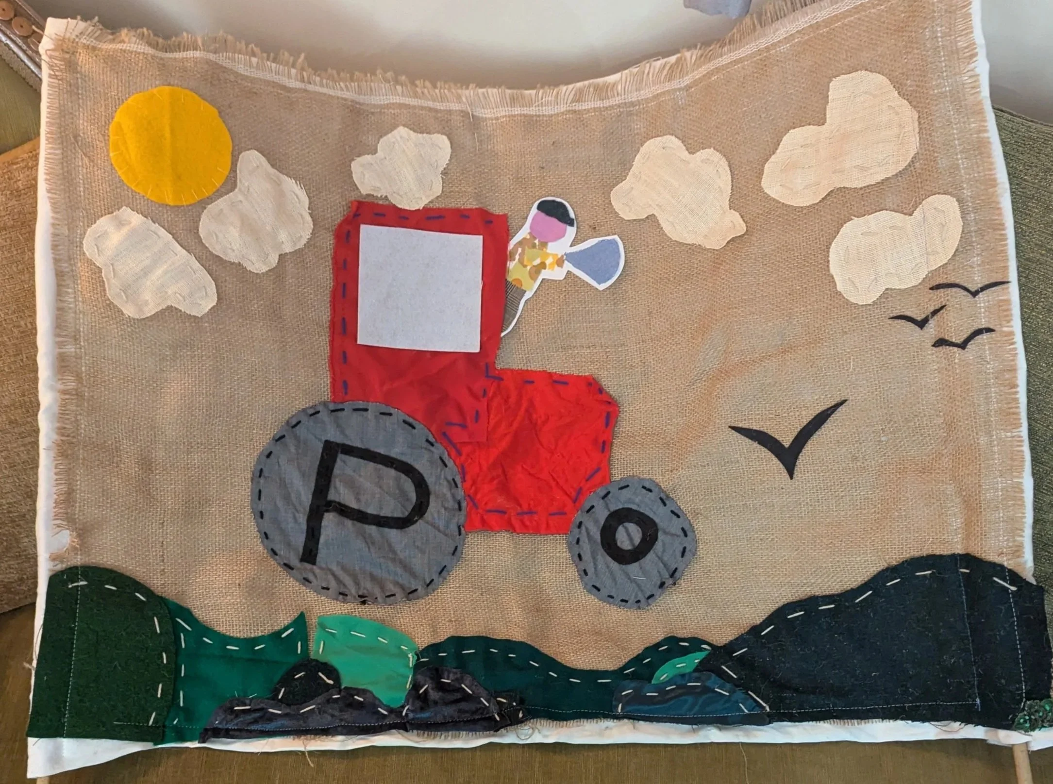 Folk art fabric scene depicting a red tractor with large gray wheels, marked with the letter P, moving through green hills with black rocks, under a light tan sky with clouds, a yellow sun, and birds.