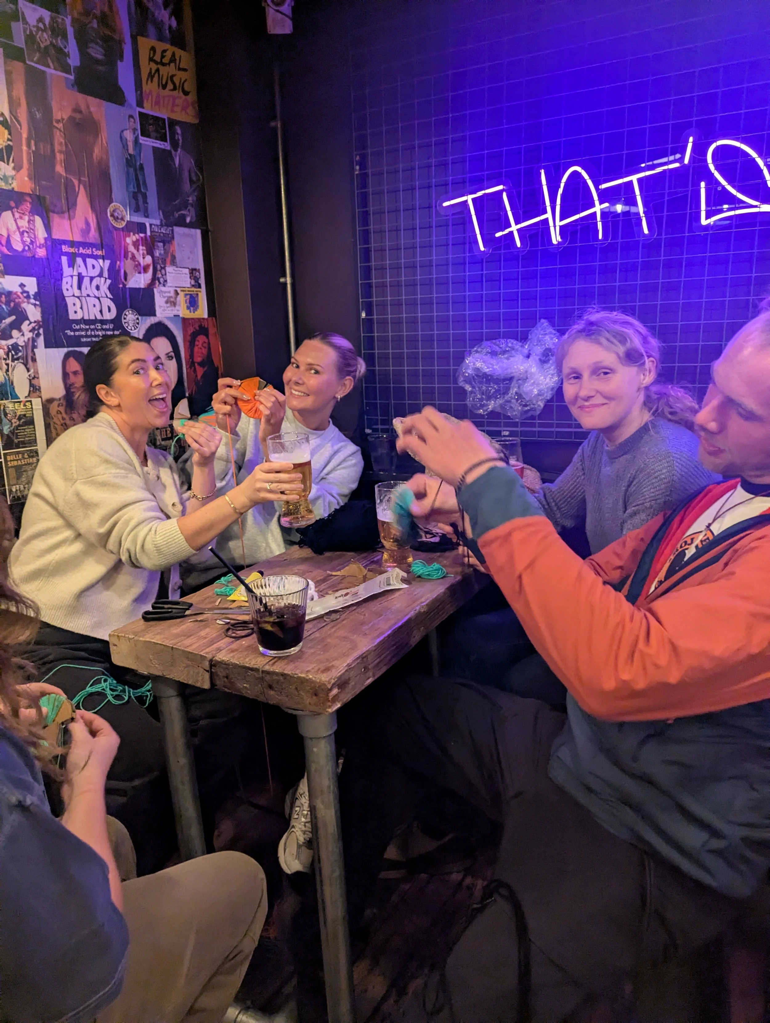 Group of friends sitting at a wooden table in a dimly lit bar with purple neon signs and wall posters, engaging in knitting and holding drinks, smiling and enjoying each other's company.