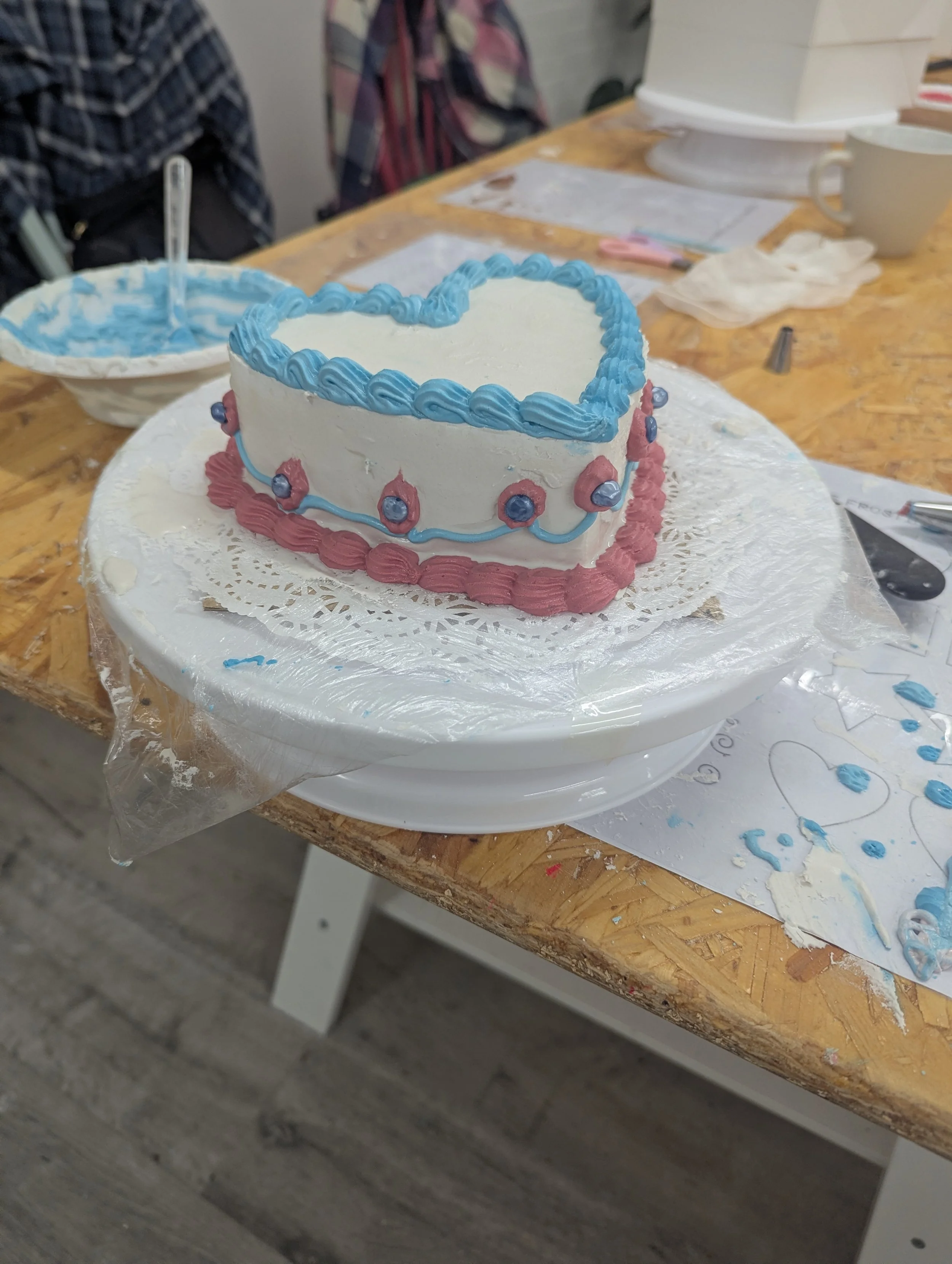 Heart-shaped birthday cake with white icing, blue border, pink base, and blue decorative accents, placed on a cake stand on a wooden table.