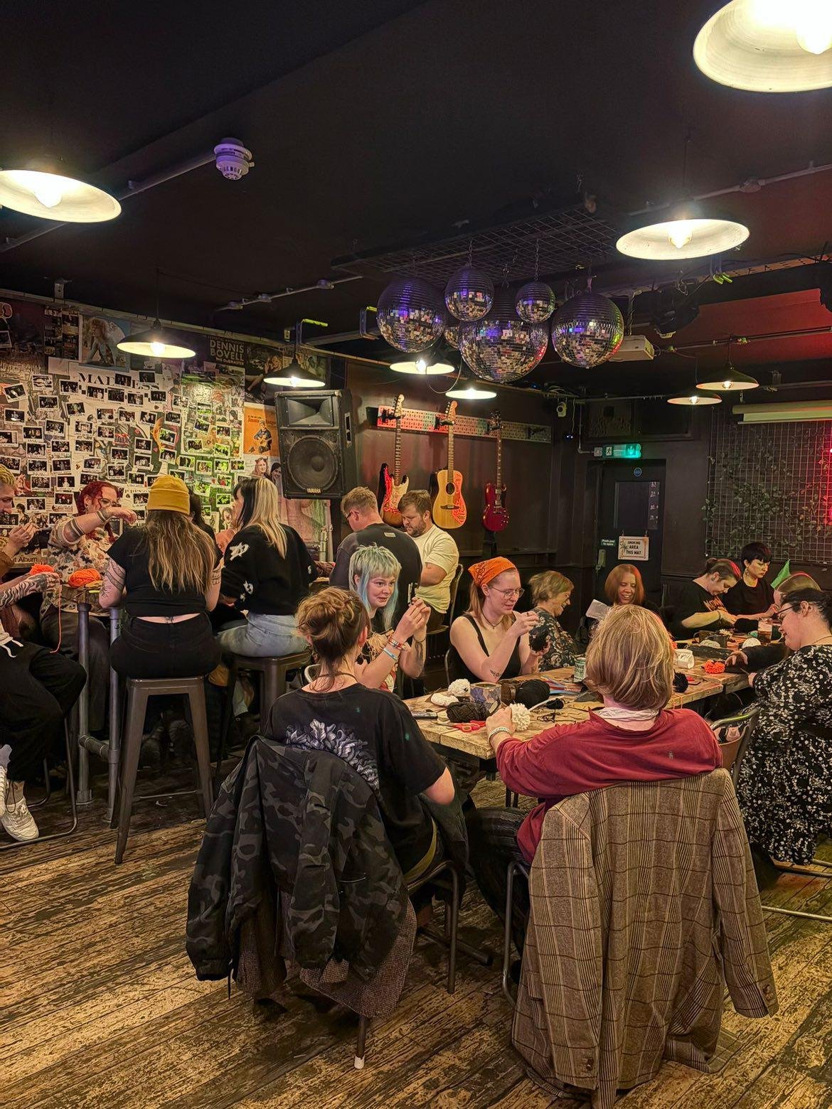 People gathered in a cozy, dimly lit music venue or bar, sitting around tables crafting pompom projects, with guitars on the wall and disco balls hanging from the ceiling.