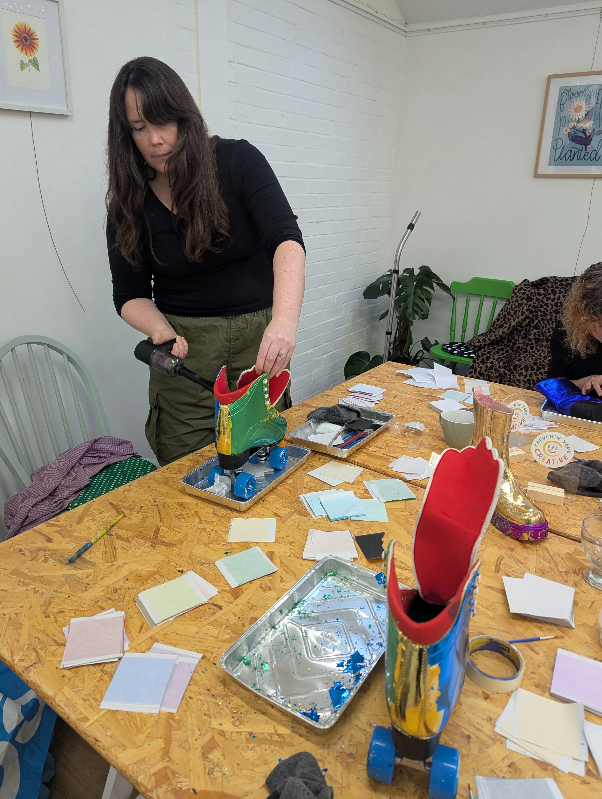 Person shaping a recycled roller skate shoe sculpture on a table surrounded by craft supplies, paper, glue, and brushes.
