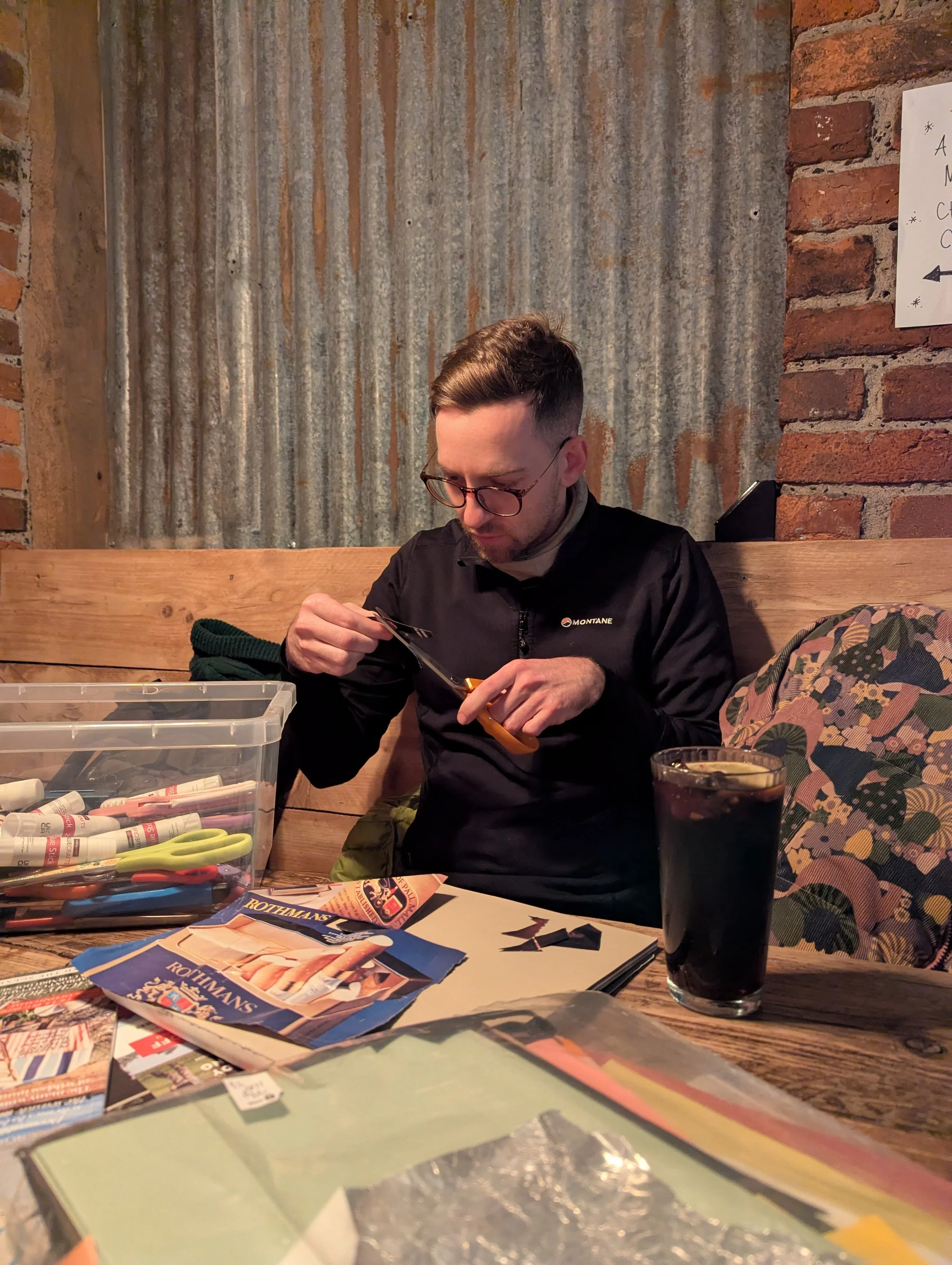 A man sitting at a wooden table, using scissors to cut paper, with craft supplies and a glass of dark soda on the table, in a rustic indoor setting.