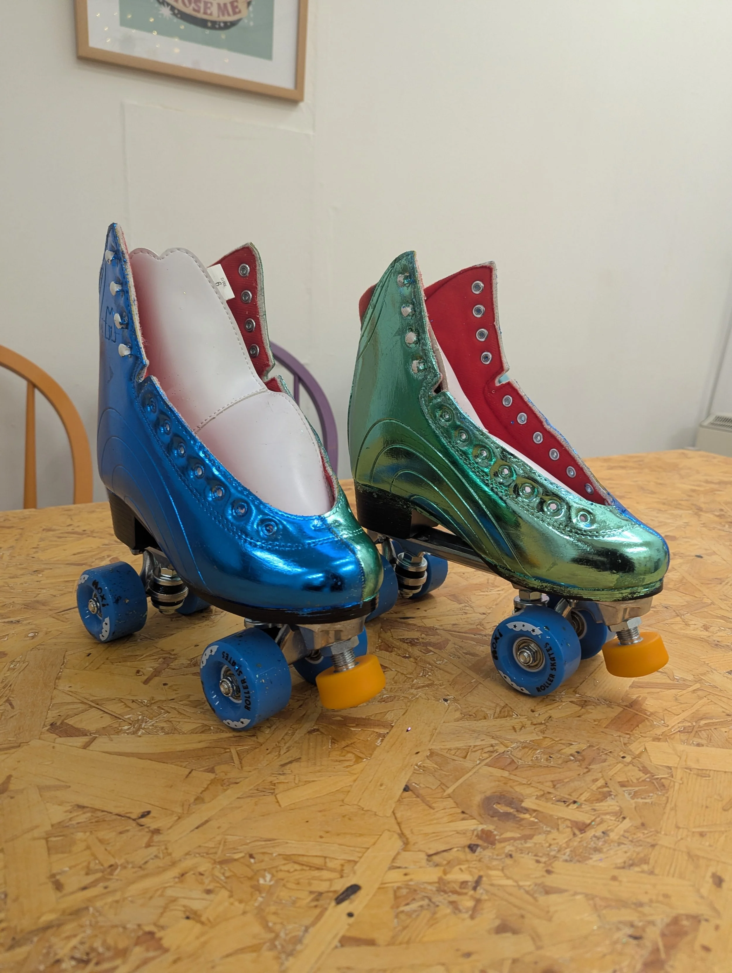 A pair of vintage roller skates with metallic blue and green shells, white and red inside linings, silver eyelets, and black wheels with yellow and blue wheels on a wooden surface.