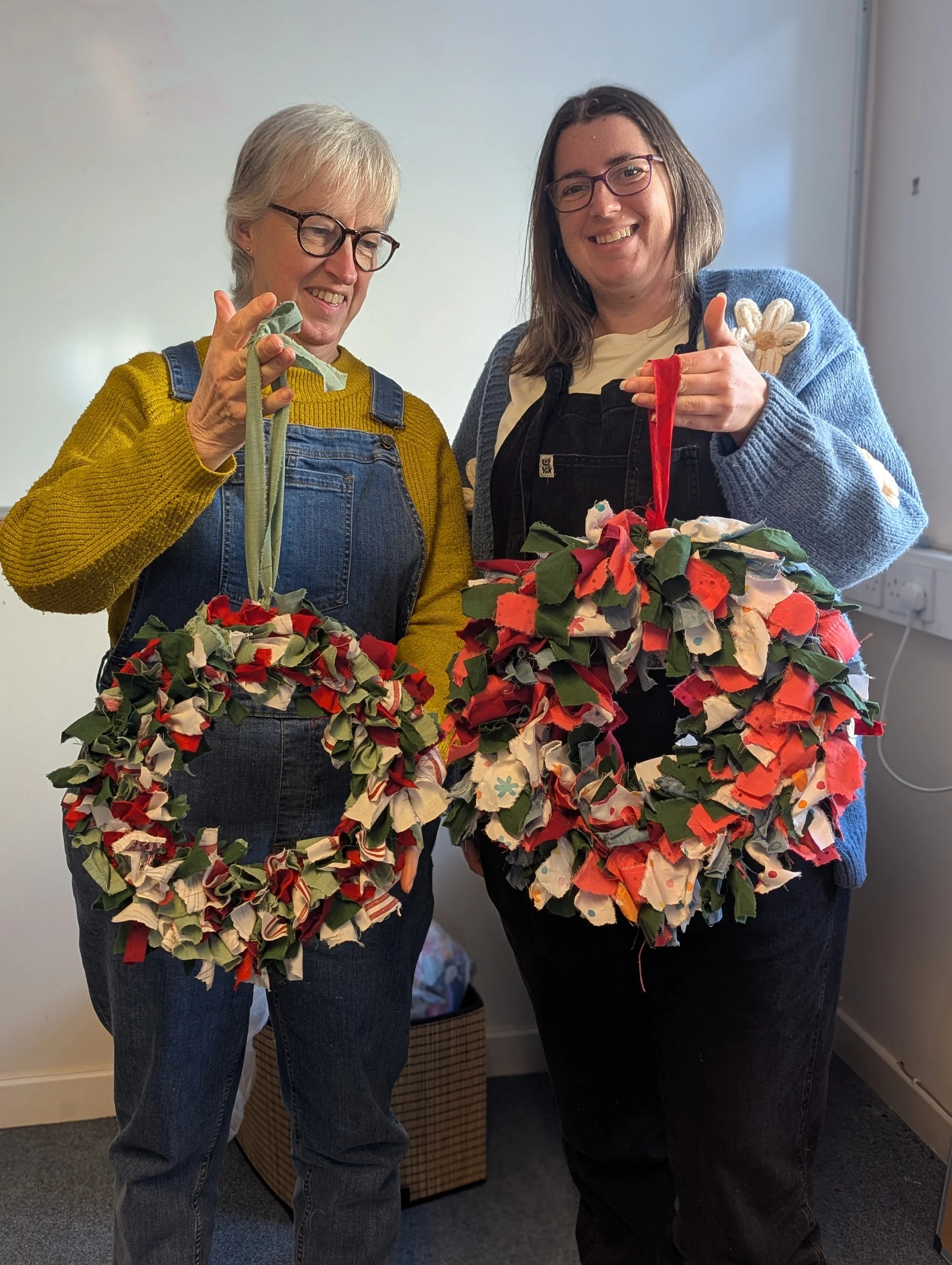 Two women smiling and holding decorated fabric wreaths made of colorful fabric scraps, standing indoors in front of a white wall.