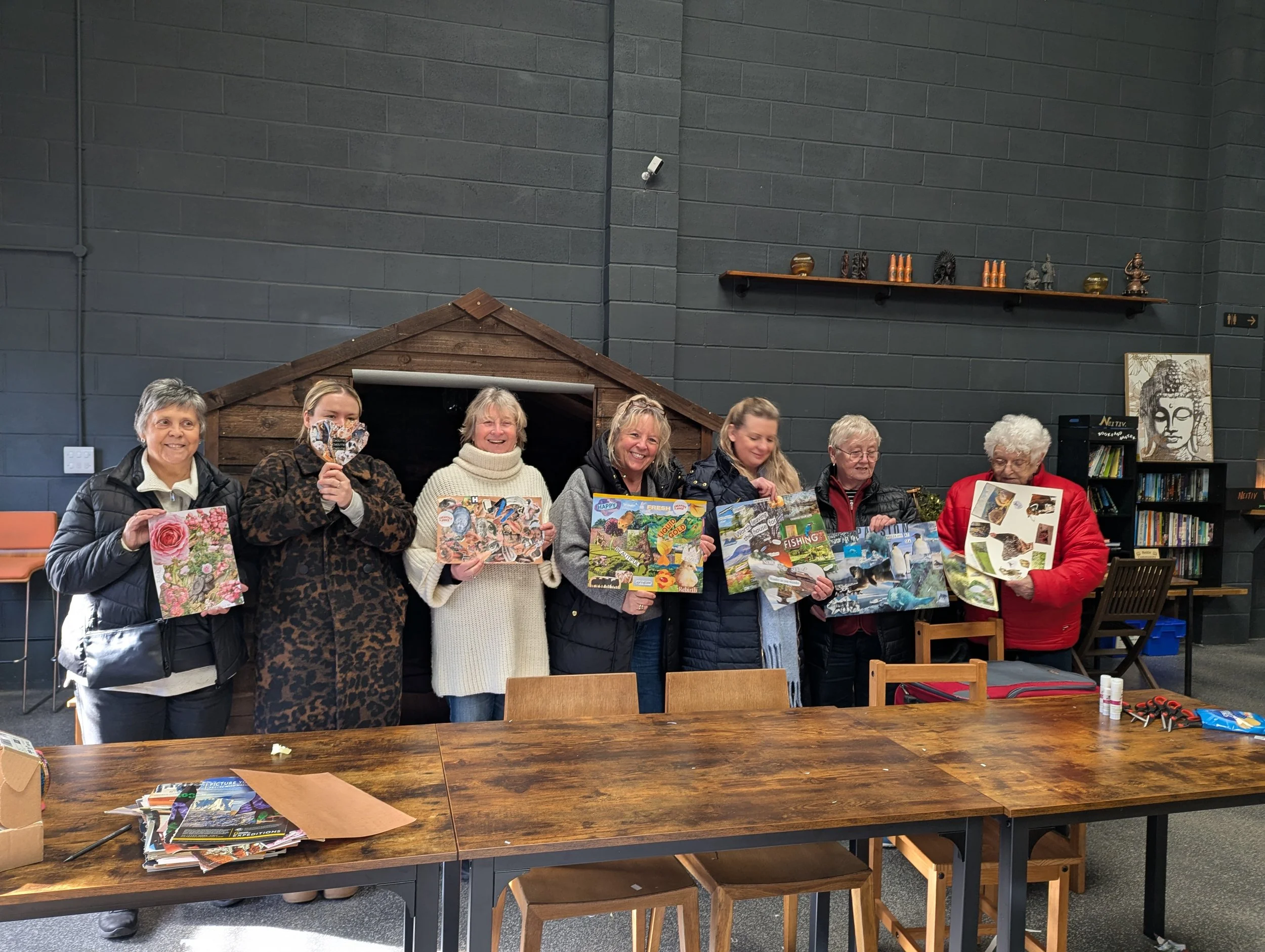 Seven women standing indoors, holding board games and puzzles, smiling. The room has dark gray walls, a wooden structure in the background, and a bookshelf with books and artwork.