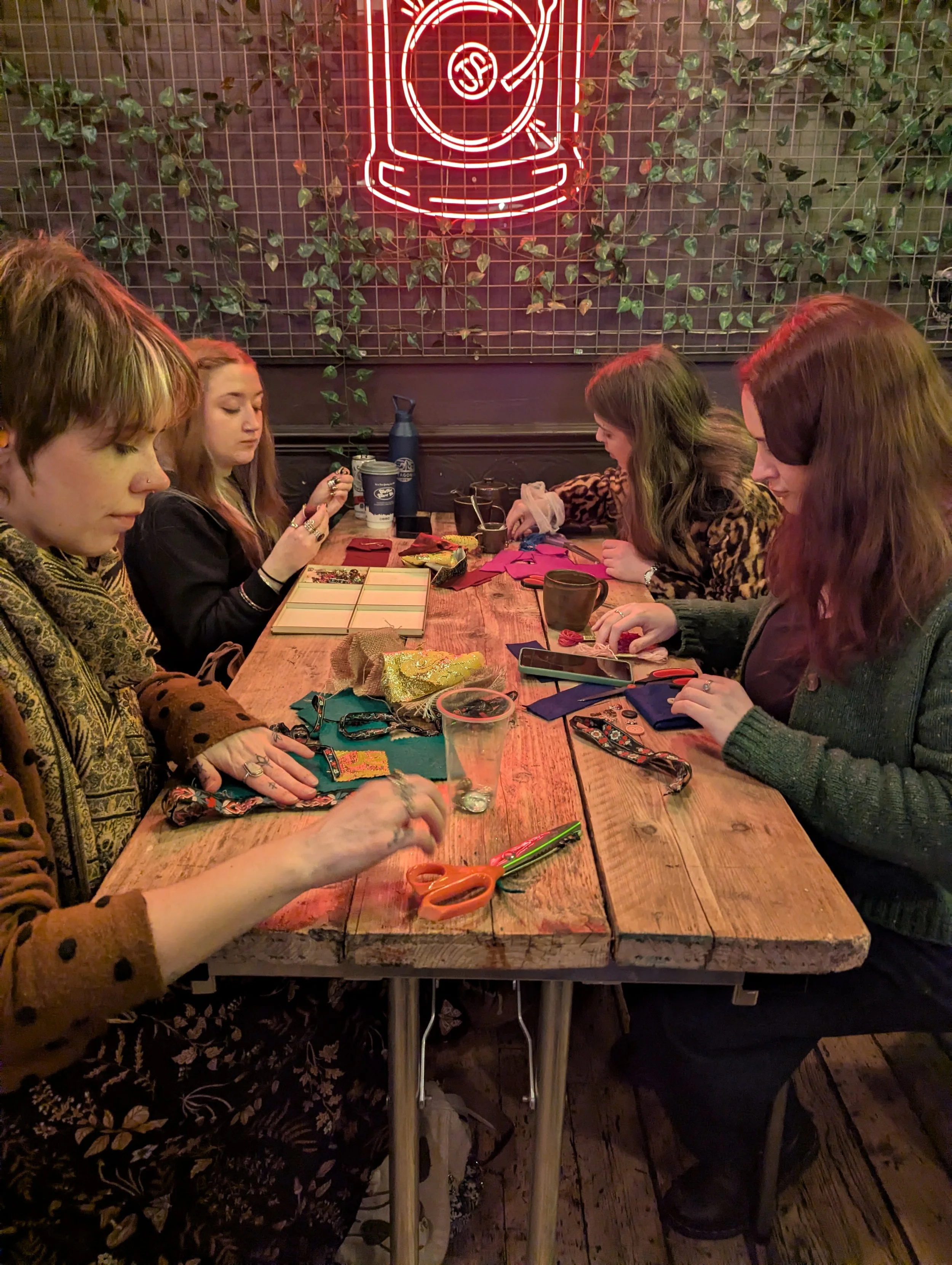 Four women sitting at a wooden table engaging in crafting activities in a cozy, dimly lit space with a neon sign of a coffee cup on a grid wall behind them and greenery.