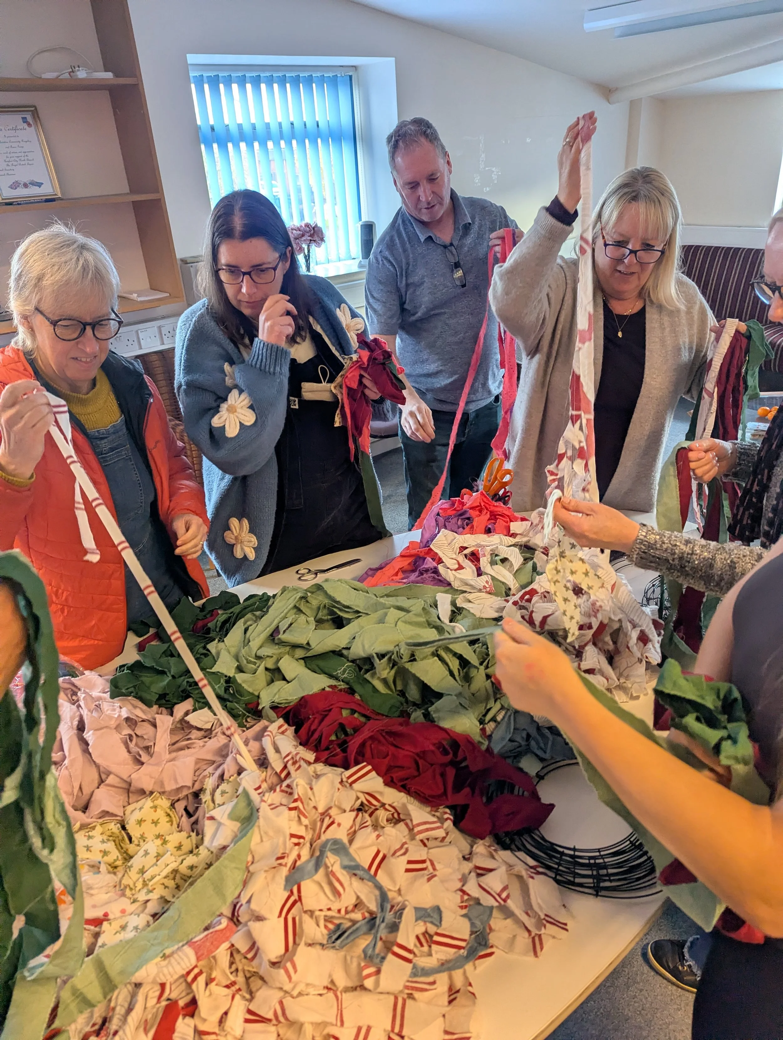 Group of people making fabric wreaths using strips of cloth at a table in a room with a window and shelves.