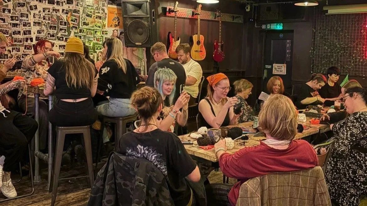 People gathered in a music-themed bar or cafe, sitting at tables and bar stools, making pompoms with yarn, guitars hanging on the wall, and photos decorating the wall.
