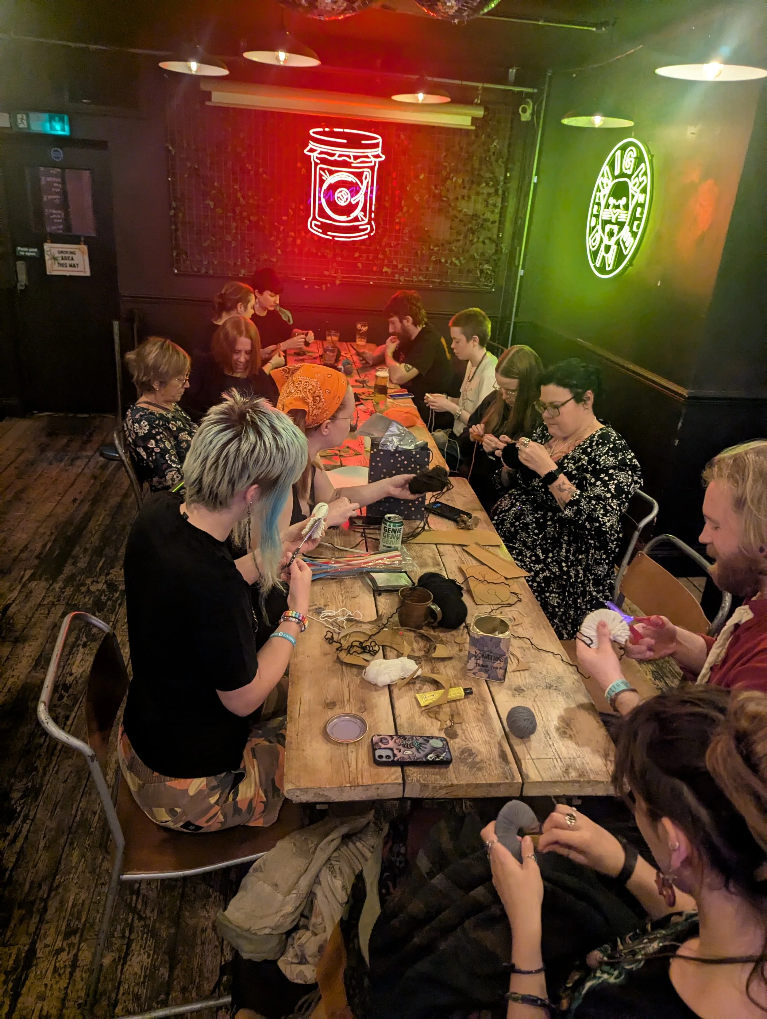 A group of people sitting around a long wooden table in a dimly lit room, engaging in craft activities like knitting or crocheting. The room has black walls with neon signs, including a red sign of a candy jar and a green sign with a circular logo. T