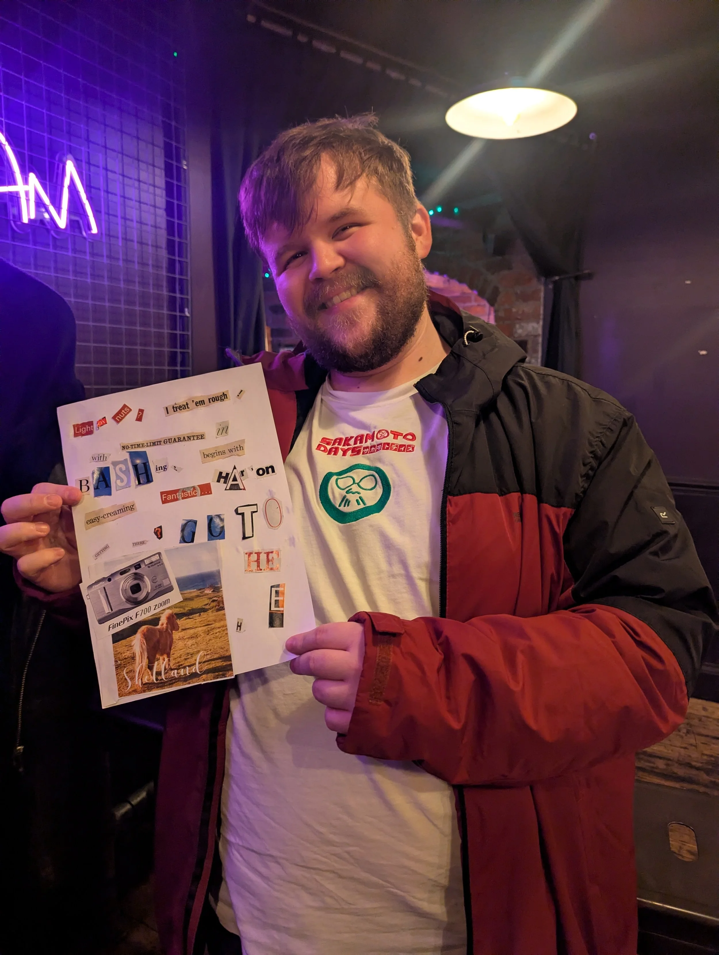 A smiling man with a beard holding a collage of cut-out words and images, standing in a dimly lit room with a neon sign.