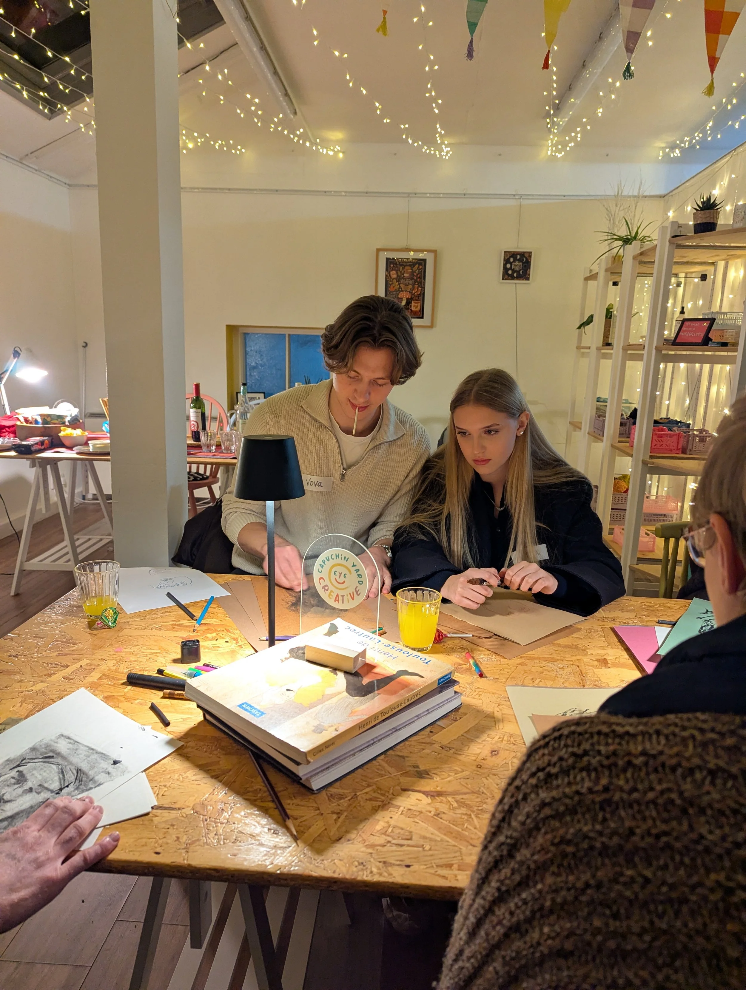 People gathered around a wooden table engaged in an art or craft activity, with art supplies and books on the table, decorated with string lights and bunting hanging from the ceiling.