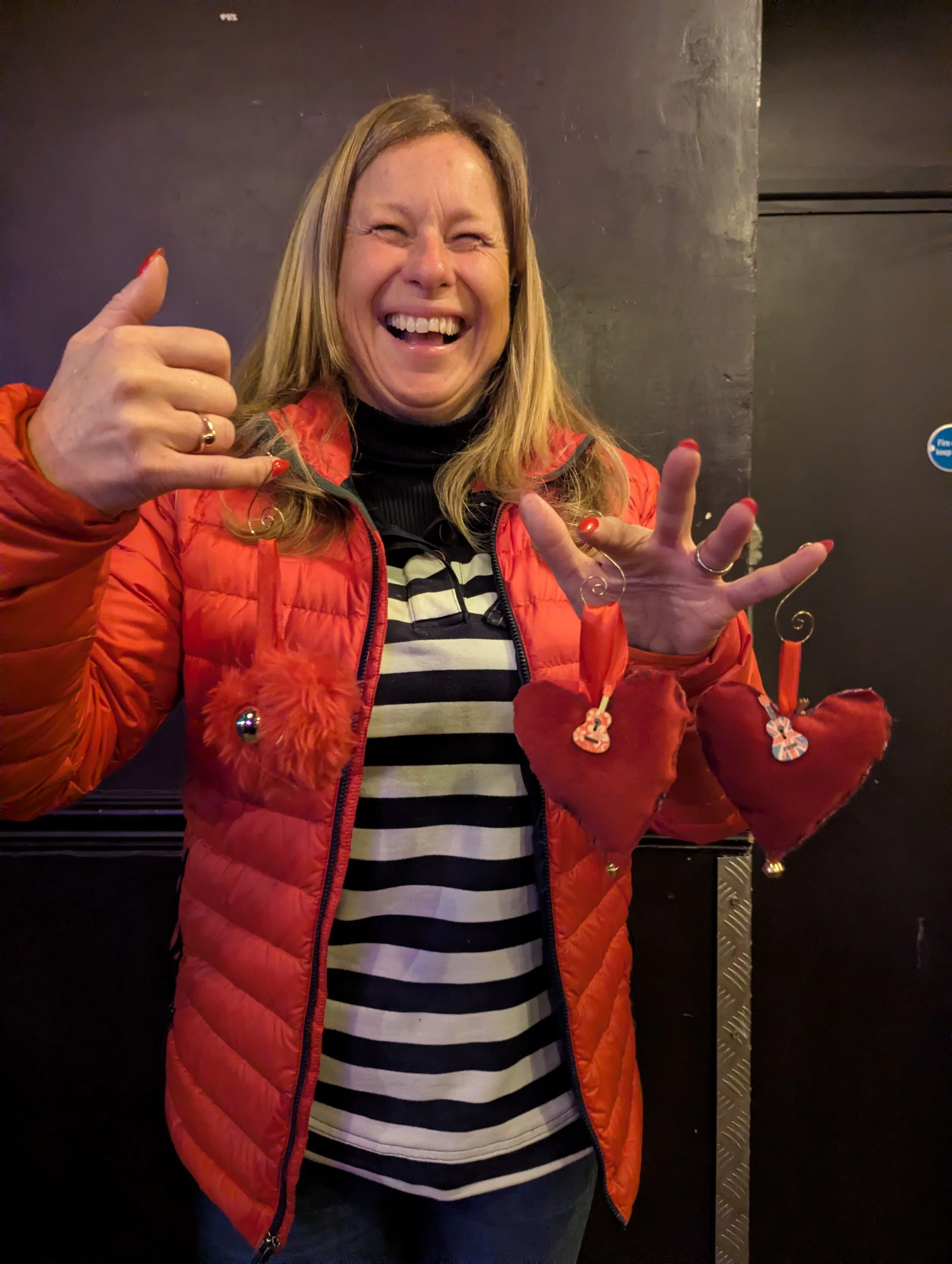 A woman smiling and making a shaka sign with her right hand, wearing a red puffy jacket with heart-shaped ornaments hanging from her sleeves, a black and white striped shirt, and standing in front of a dark wall.