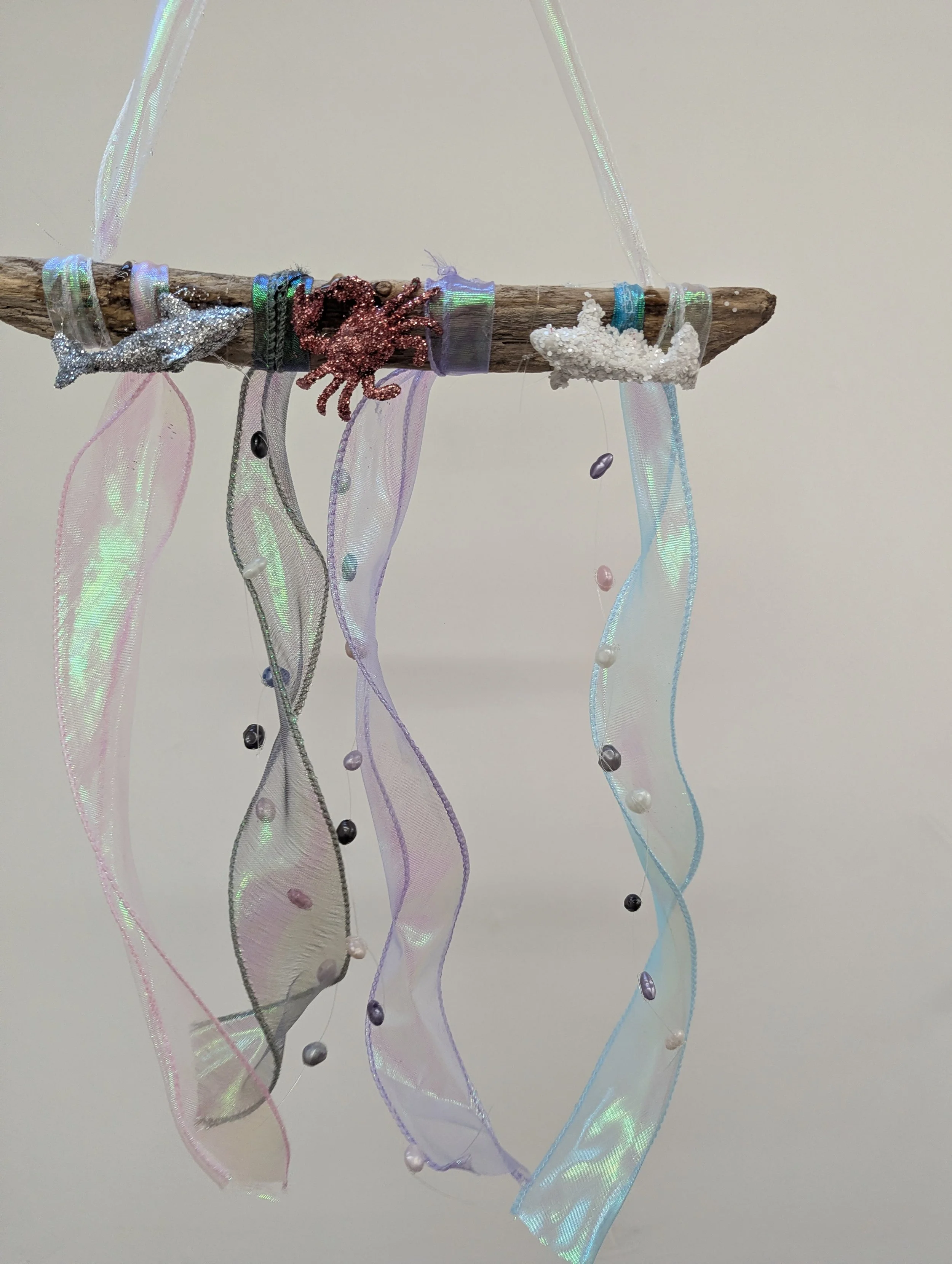 Decorative hanging craft with a driftwood stick, colorful ribbons, beaded strings, and small sea creature and starfish embellishments.
