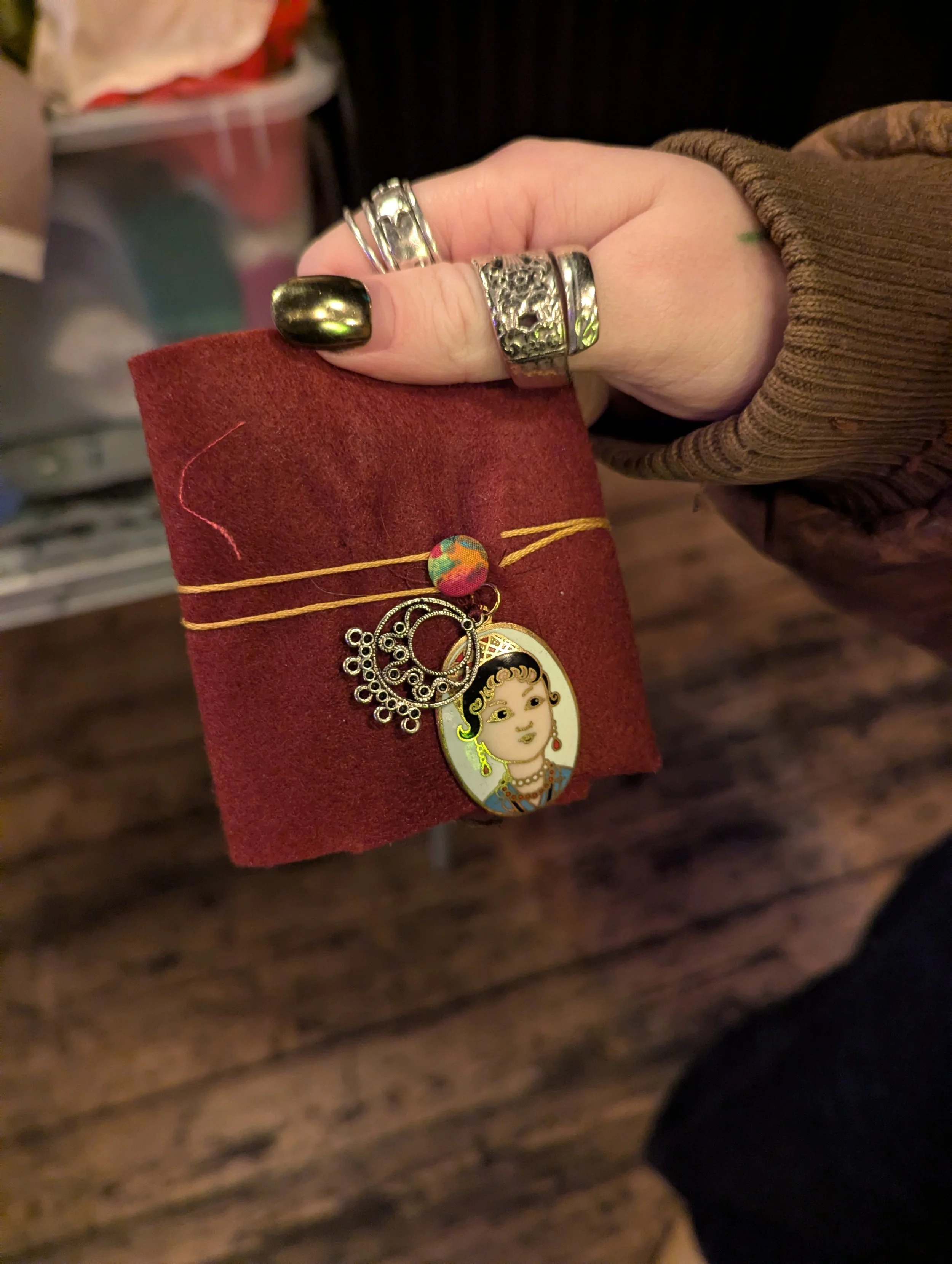 Hand holding a red jewelry roll with rings and a bracelet featuring a woman’s portrait. The hand is adorned with multiple silver rings and painted dark metallic nails. The background shows a wooden floor and some out-of-focus storage.