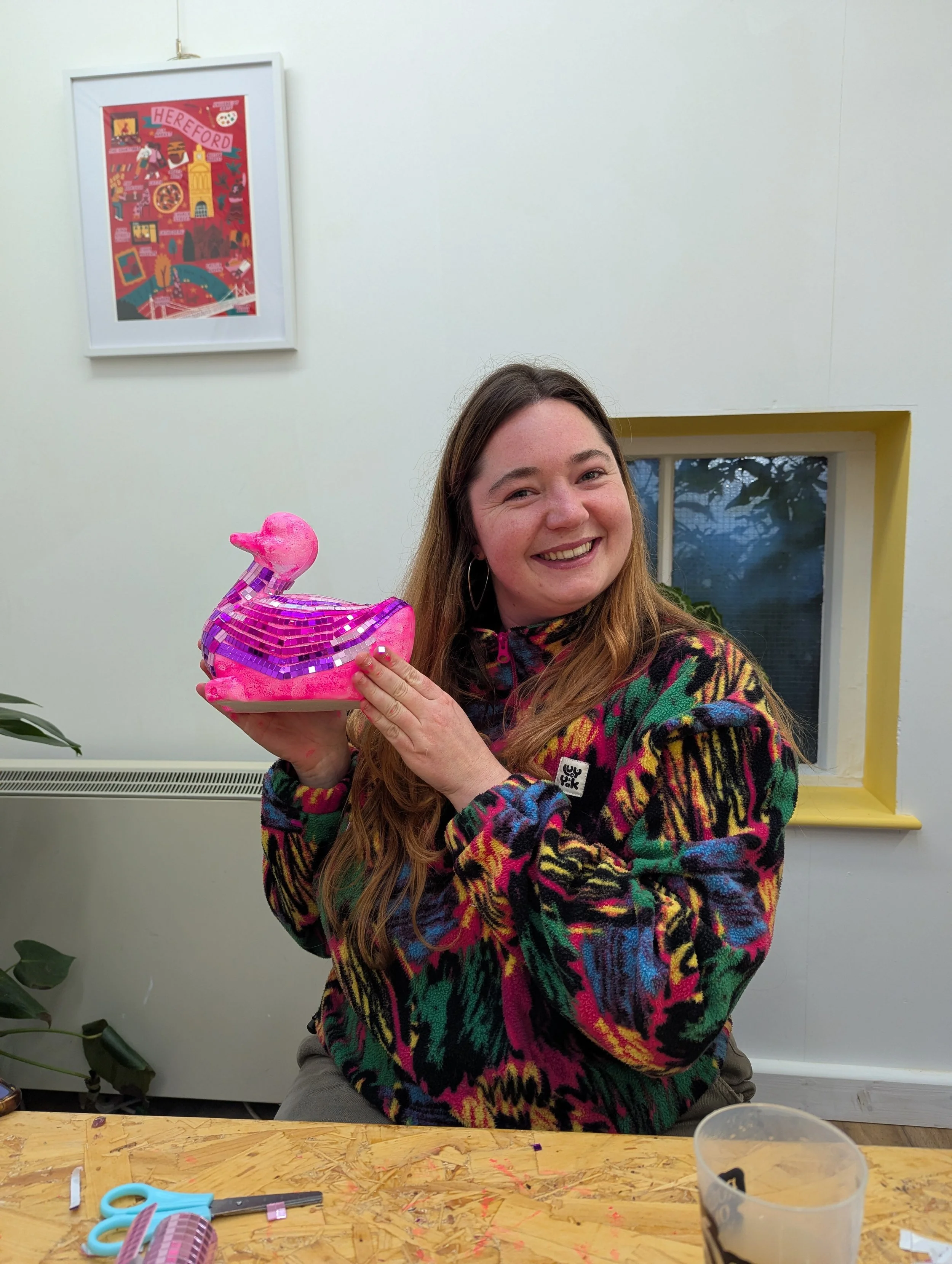 A woman with long brown hair and hoop earrings smiling and holding a pink inflatable rubber duck. She is wearing a colorful patterned fleece jacket. The background shows a white wall with a window and a framed colorful artwork.
