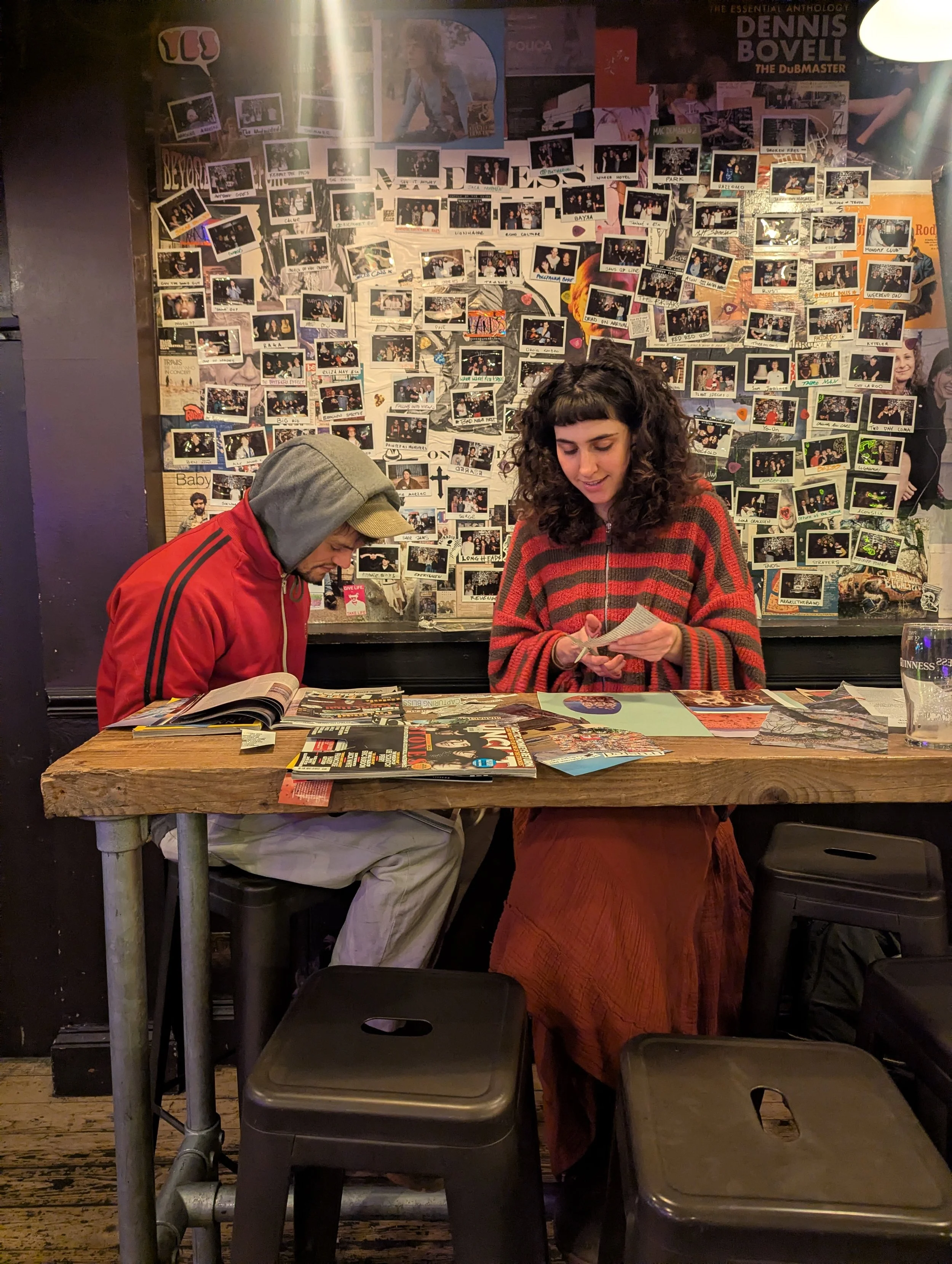 A man and woman sit at a rustic wooden table covered with magazines and brochures inside a café. The man, wearing a red jacket and gray hoodie, reads a magazine. The woman, in a red and black striped sweater and a long skirt, looks at a piece of pape