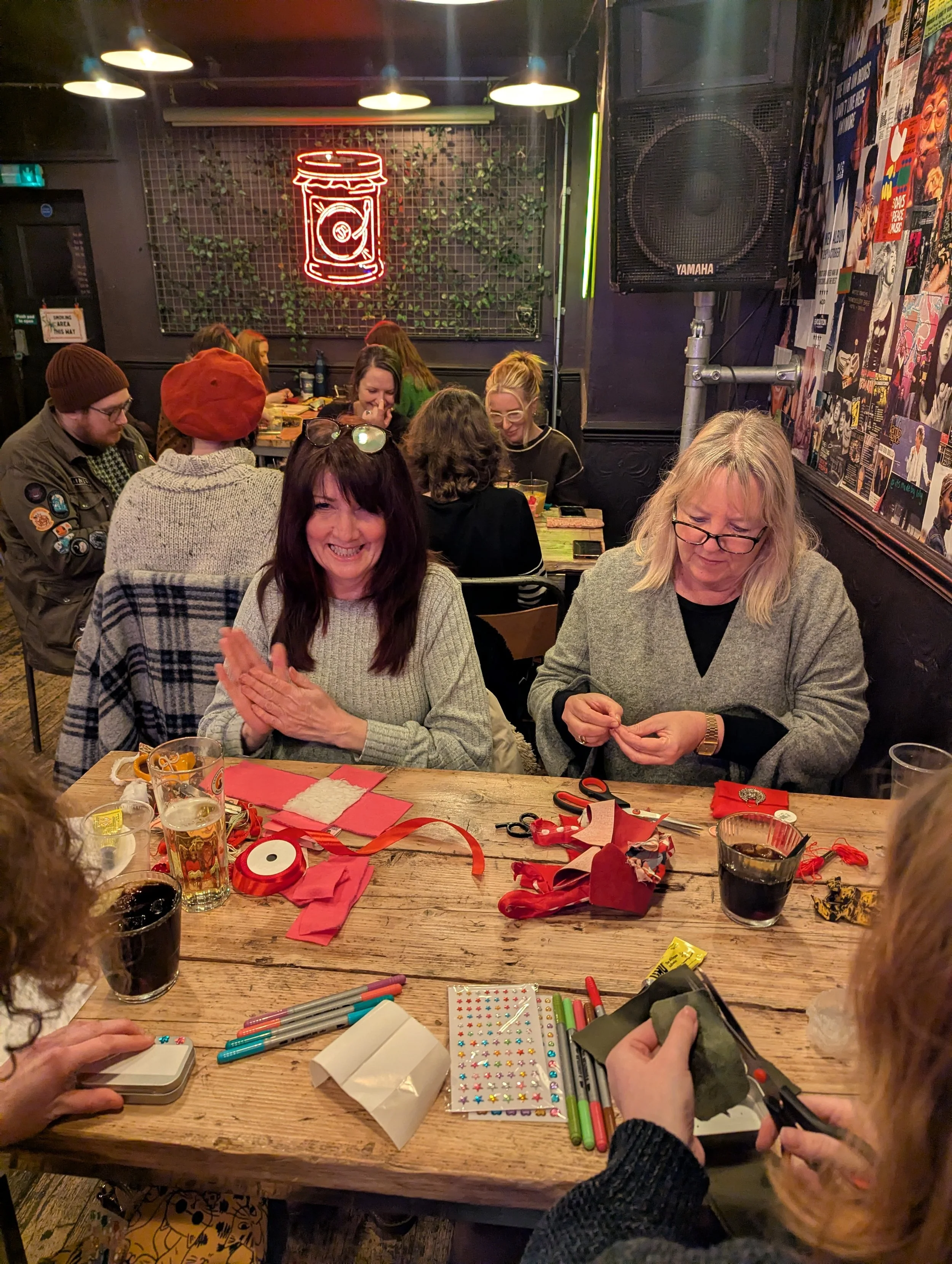 People sitting at a wooden table in a cozy, decorated restaurant or bar, engaging in arts and crafts with various supplies like scissors, ribbons, pens, and decorated stickers, with some drinks on the table, and a neon sign of a coffee cup in the bac