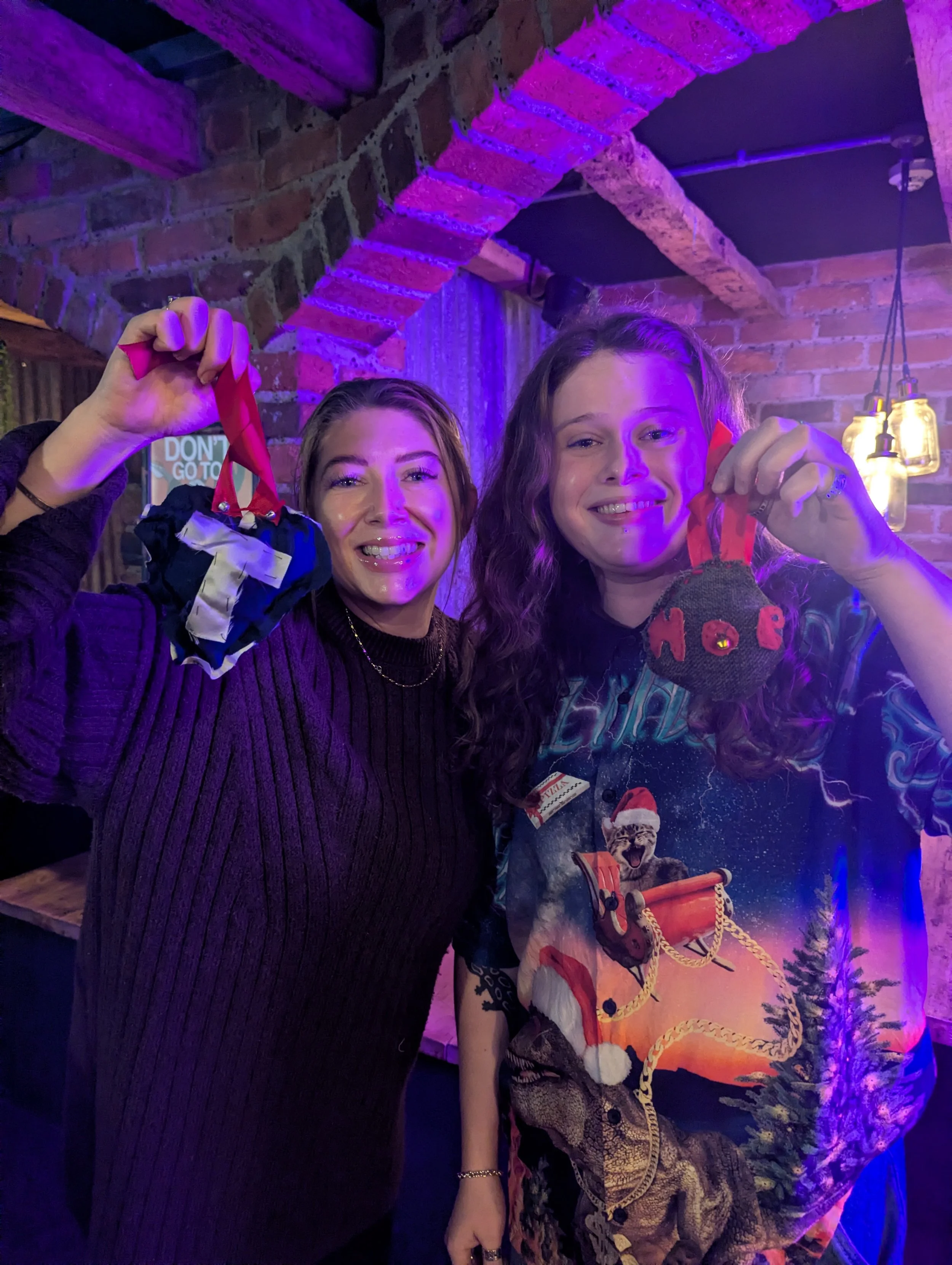 Two women standing in a brick-walled room with purple lighting, each holding a small Halloween-themed sock ornament with red ribbons, smiling at the camera.