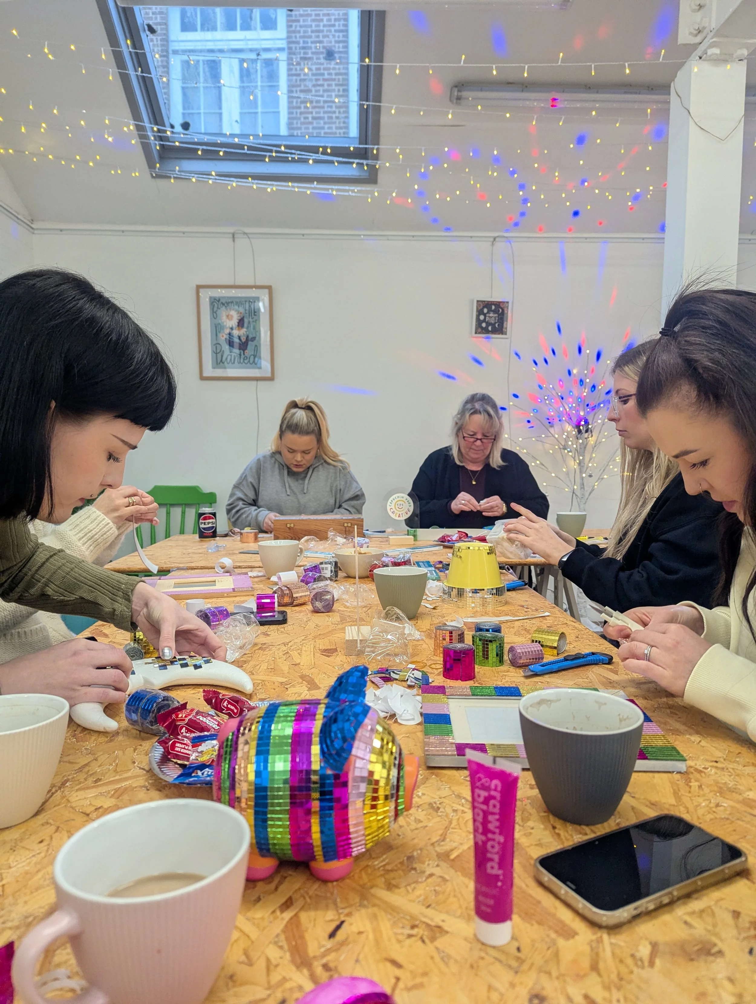 A group of women sitting around a wooden table decorating colorful items, possibly for a craft or celebration. The table has various craft supplies, cups, and snacks. The room has string lights and disco ball-like decorations on the ceiling, with a w