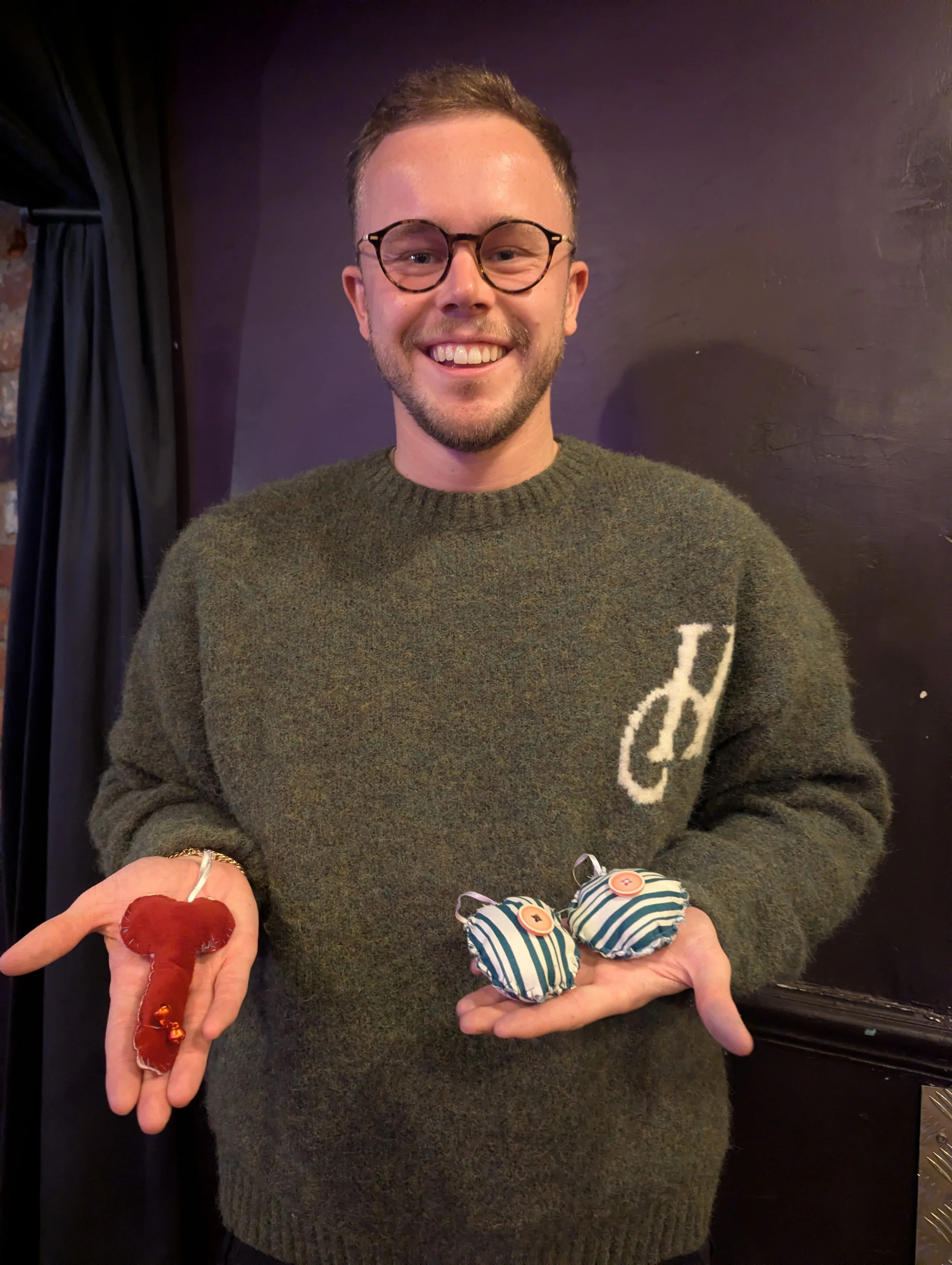 A man with glasses and a green sweater smiling while holding handmade fabric ornaments, one shaped like a red chili pepper and two striped with buttons.