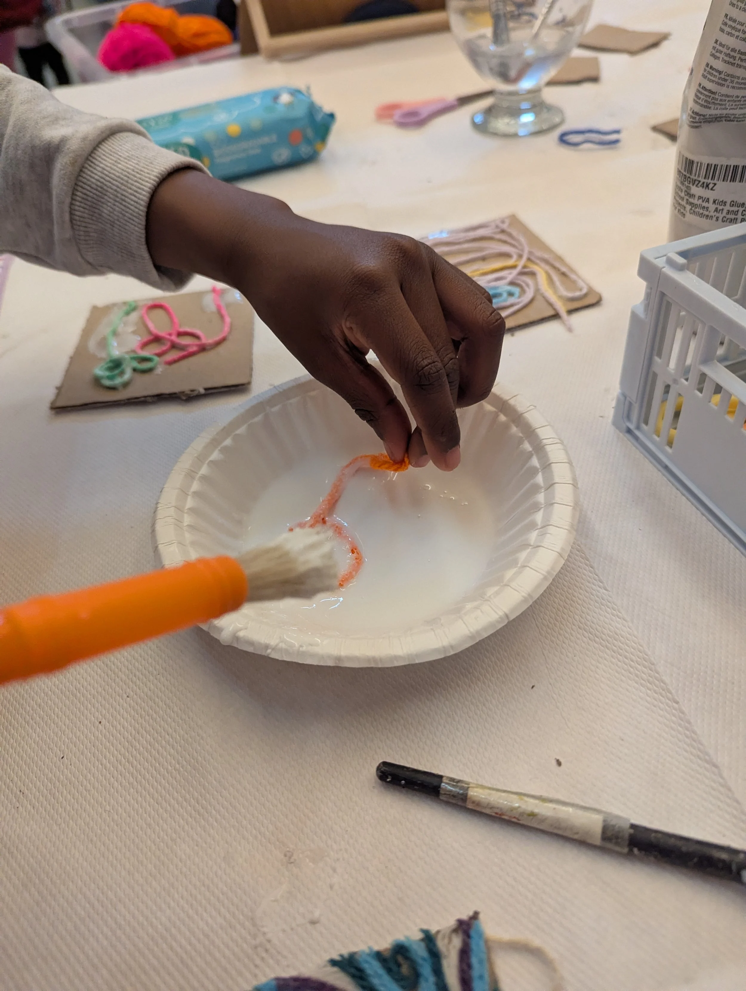 Person holding a piece of yarn over a bowl filled with a white, milky liquid, using a brush in a craft activity. Various craft supplies, including yarn, scissors, and glue, are scattered on the table.