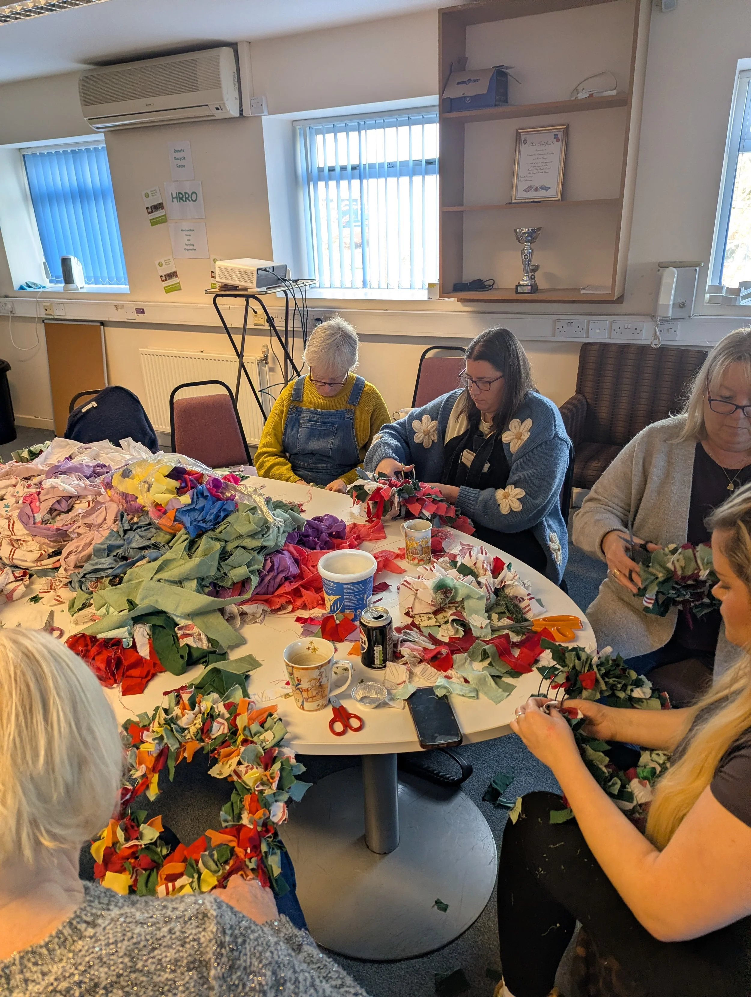 People crafting holiday wreaths with colorful fabric and supplies on a table in a well-lit room.
