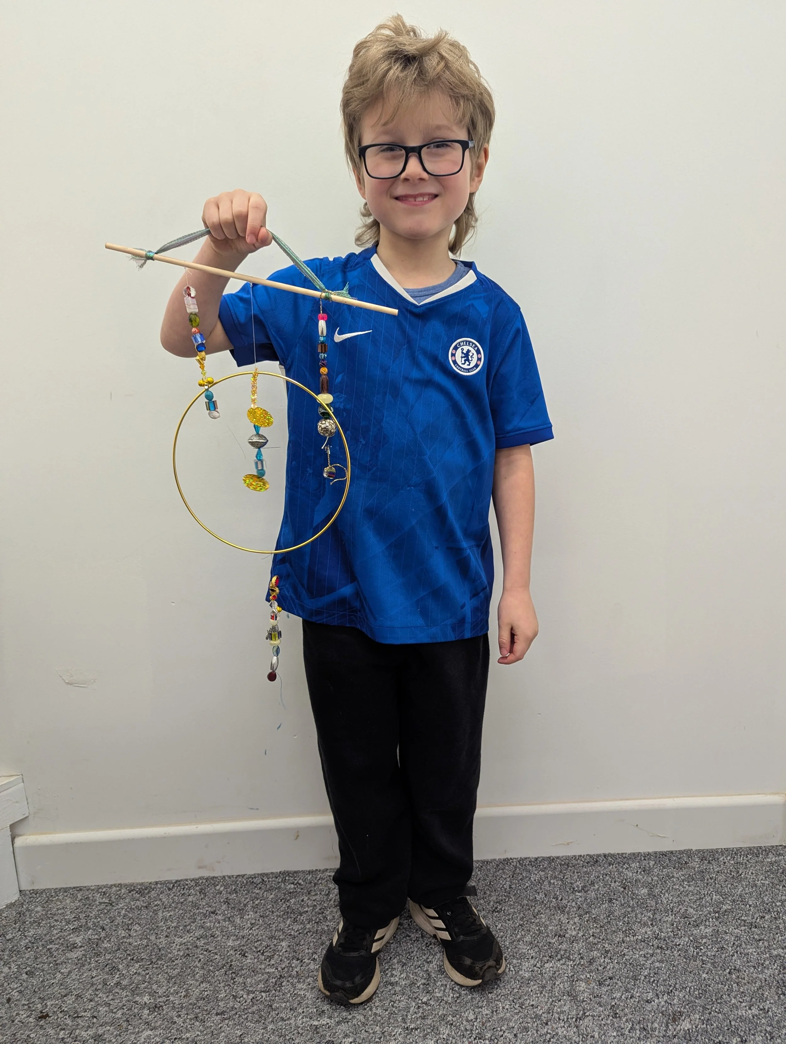 Young boy wearing glasses and a Chelsea football jersey smiling and holding a handmade craft mobile with beads and small ornaments against a plain white wall.