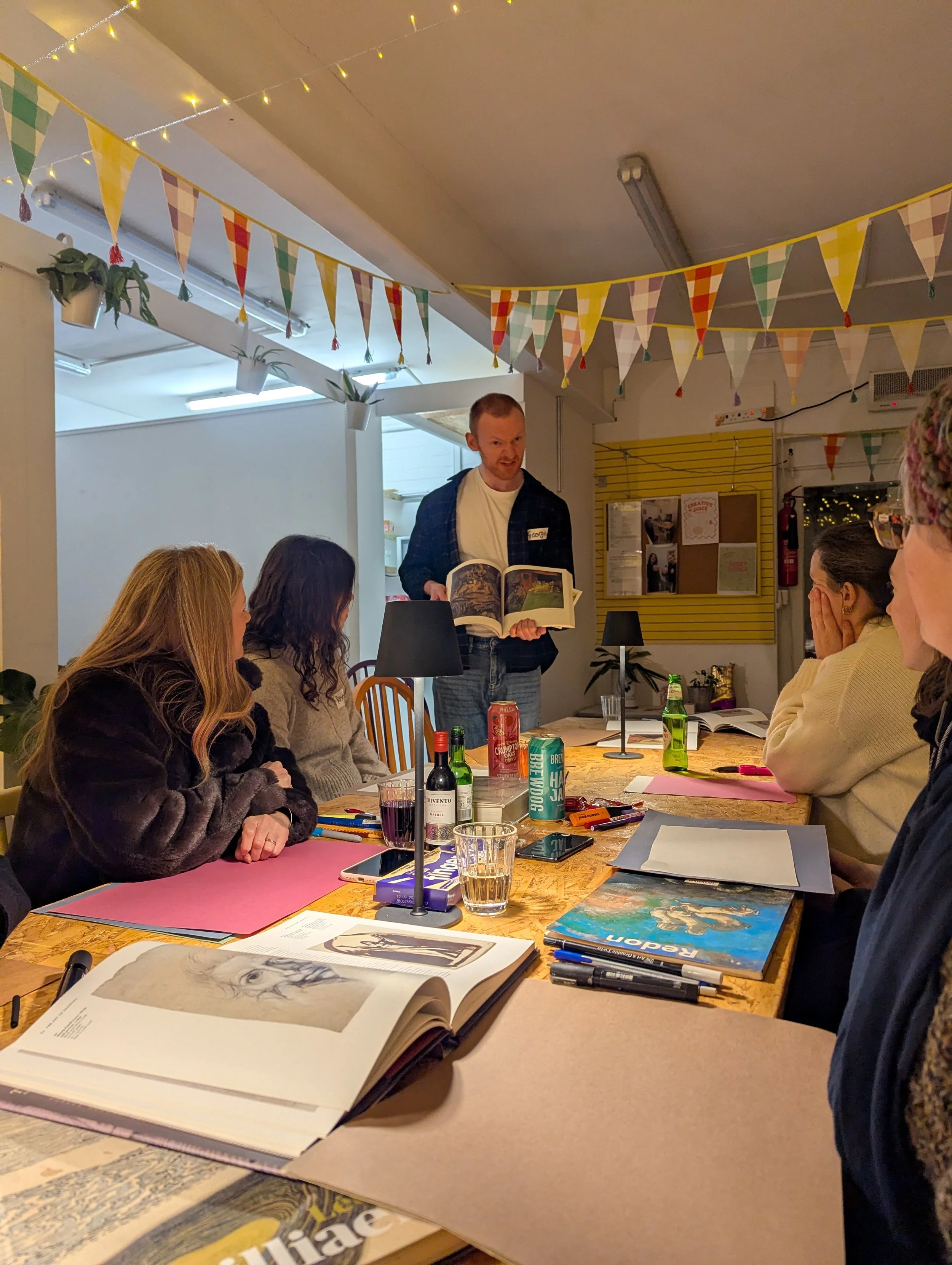 A man standing and reading from a book to a group of women seated at a wooden table, decorated with colorful triangular banners and string lights.