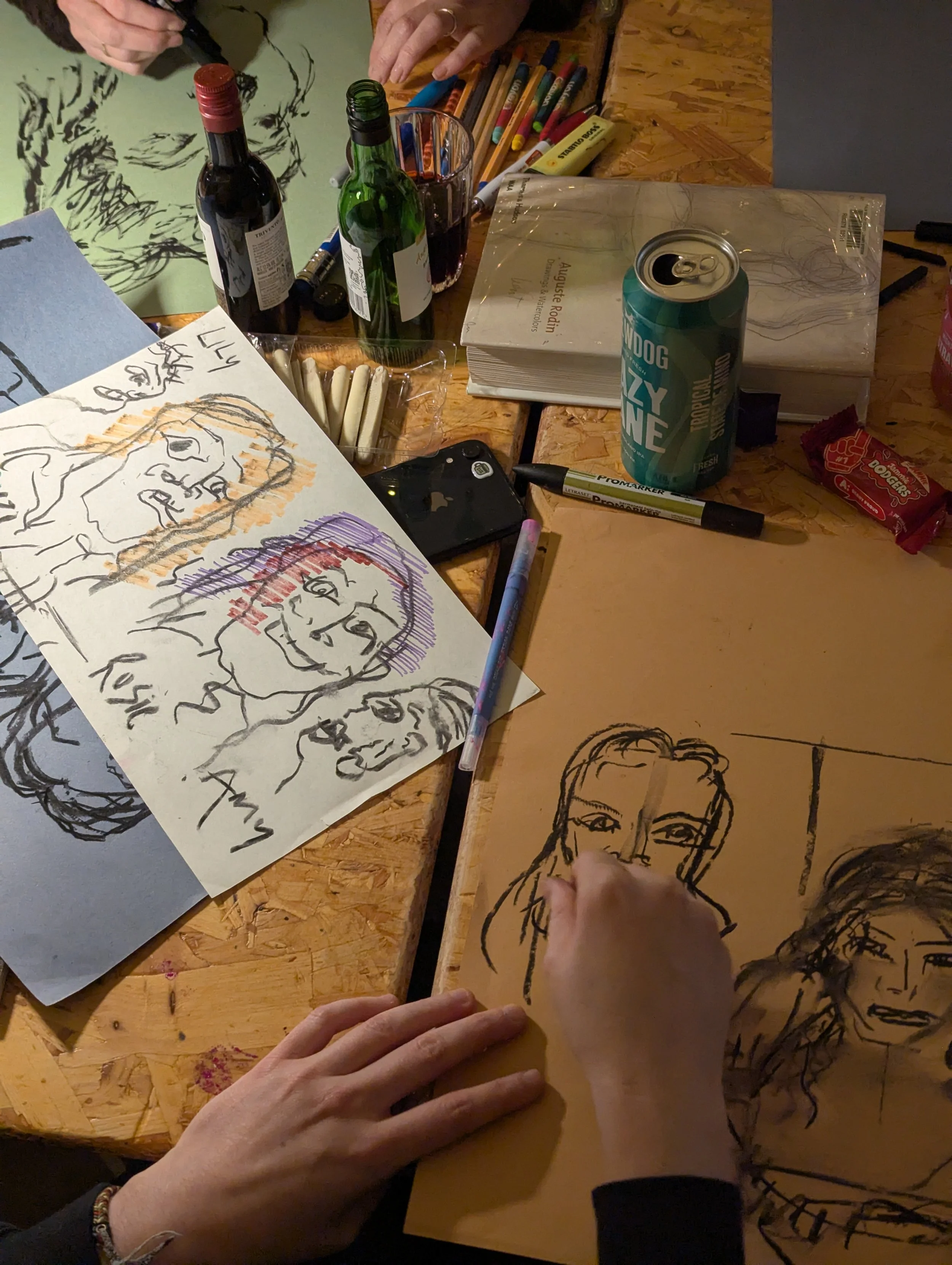 Artistic workspace with sketches of faces on paper, various drawing tools, a smartphone, bottles of wine, a can of soda, and snacks on a wooden table.