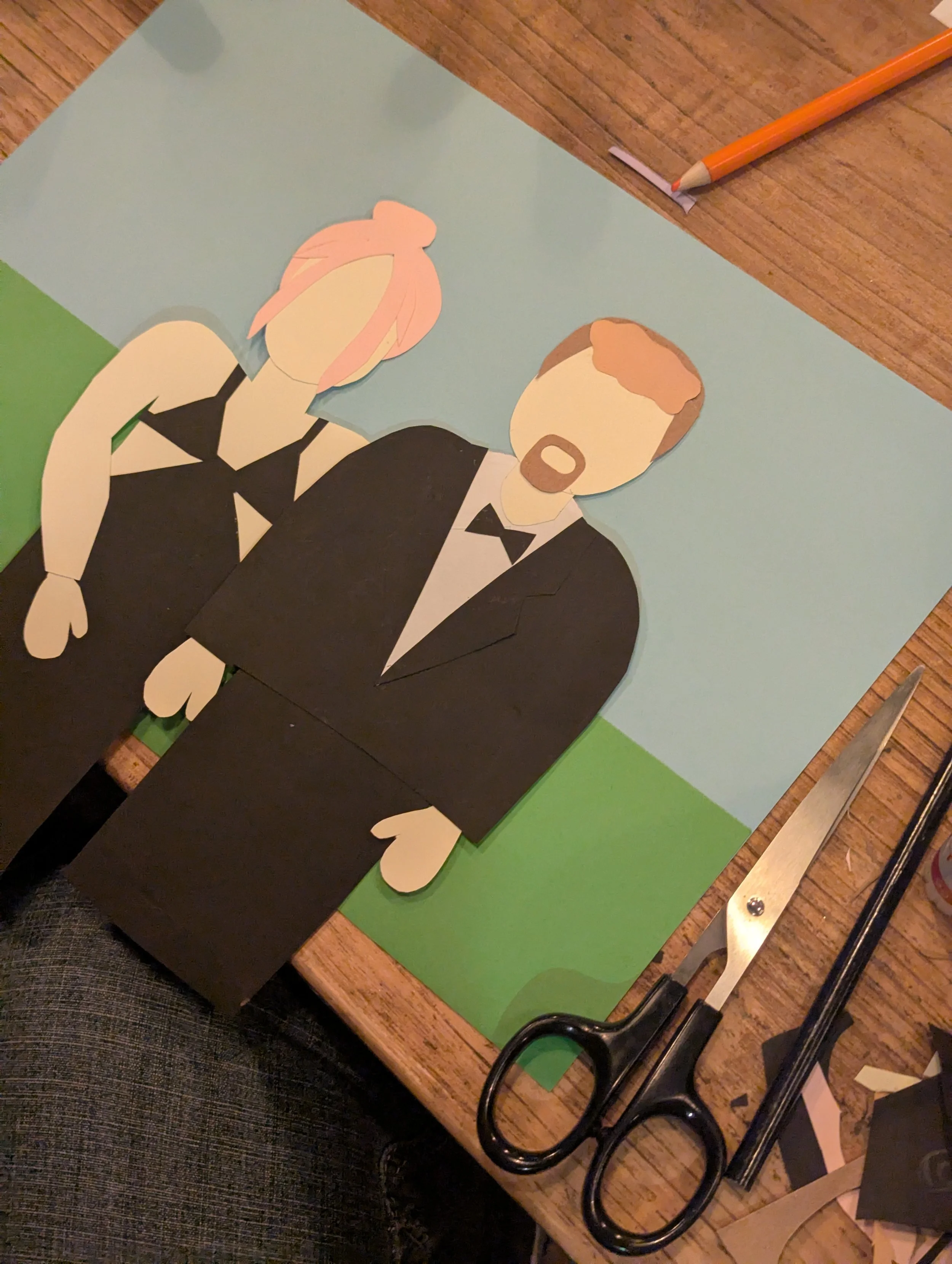 Paper cutout of a man in a tuxedo and a woman in a dress, assembled on a wooden table, with scissors, a pen, and cut paper pieces around.