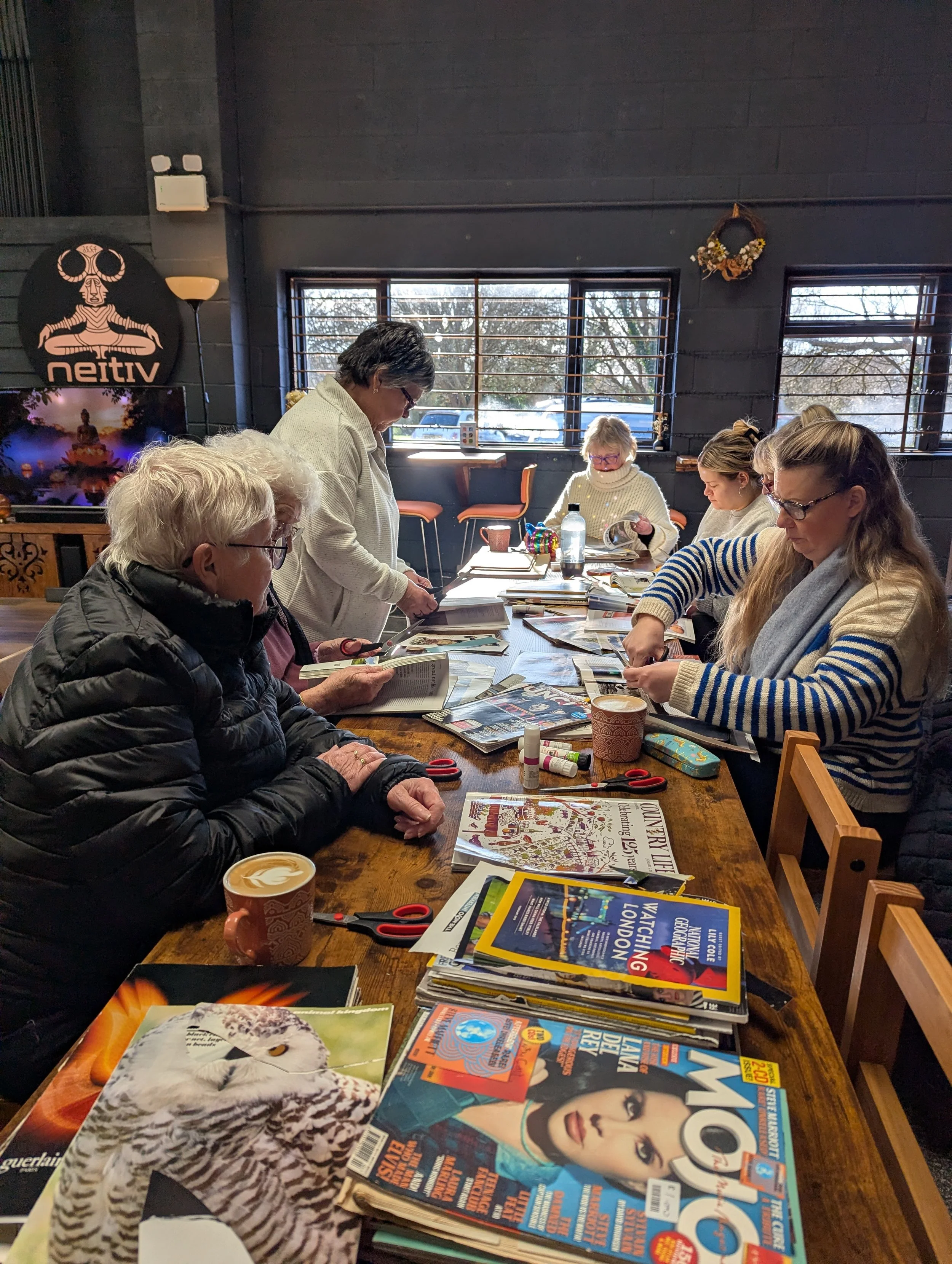 A group of six women sitting around a large wooden table, reading magazines and working on craft projects in a room with large windows, decorated with a wreath and a sign that reads 'ネイティブ' on the wall.