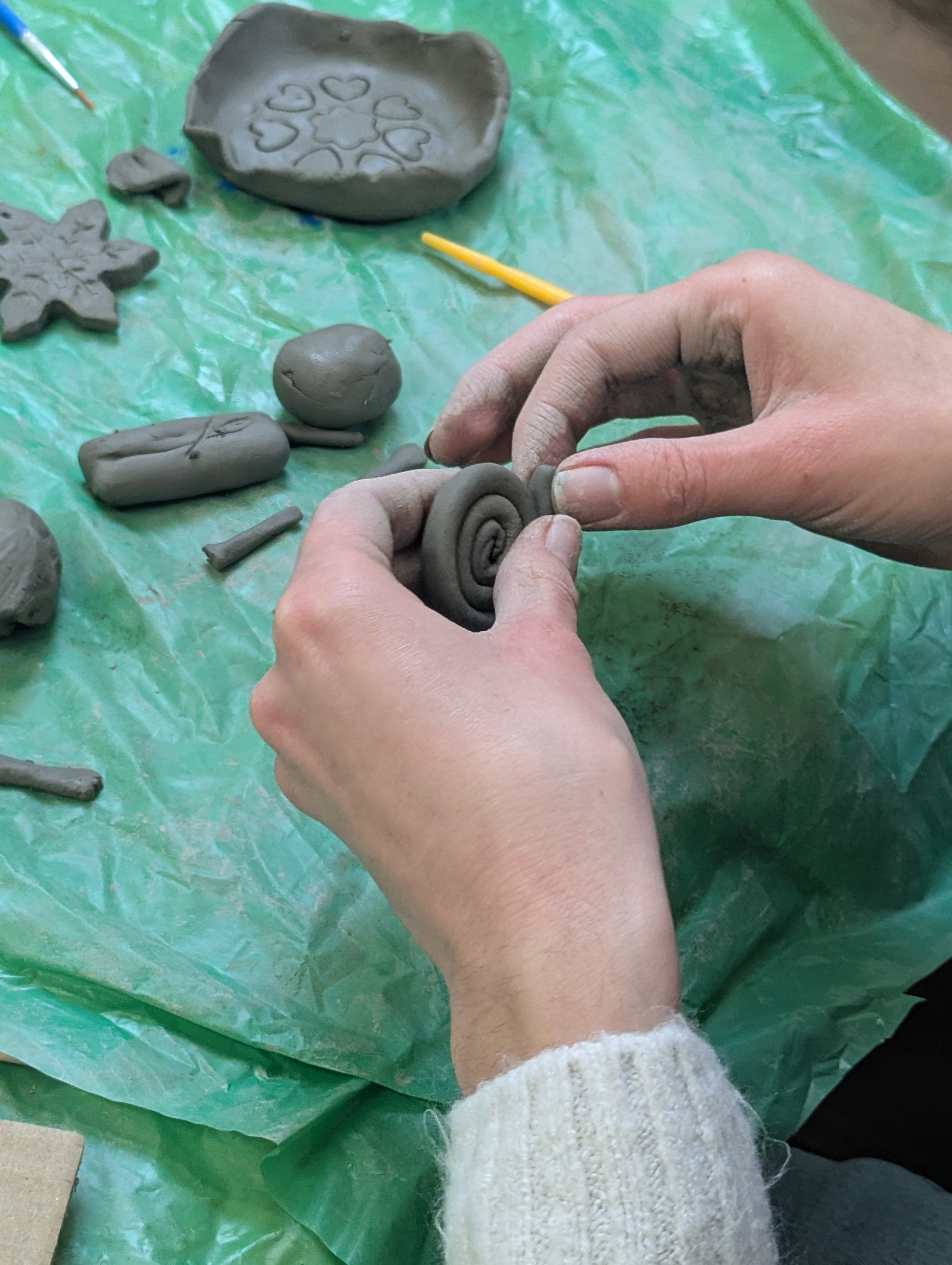 Hands shaping a spiral of gray modeling clay, surrounded by various clay creations on a green surface.