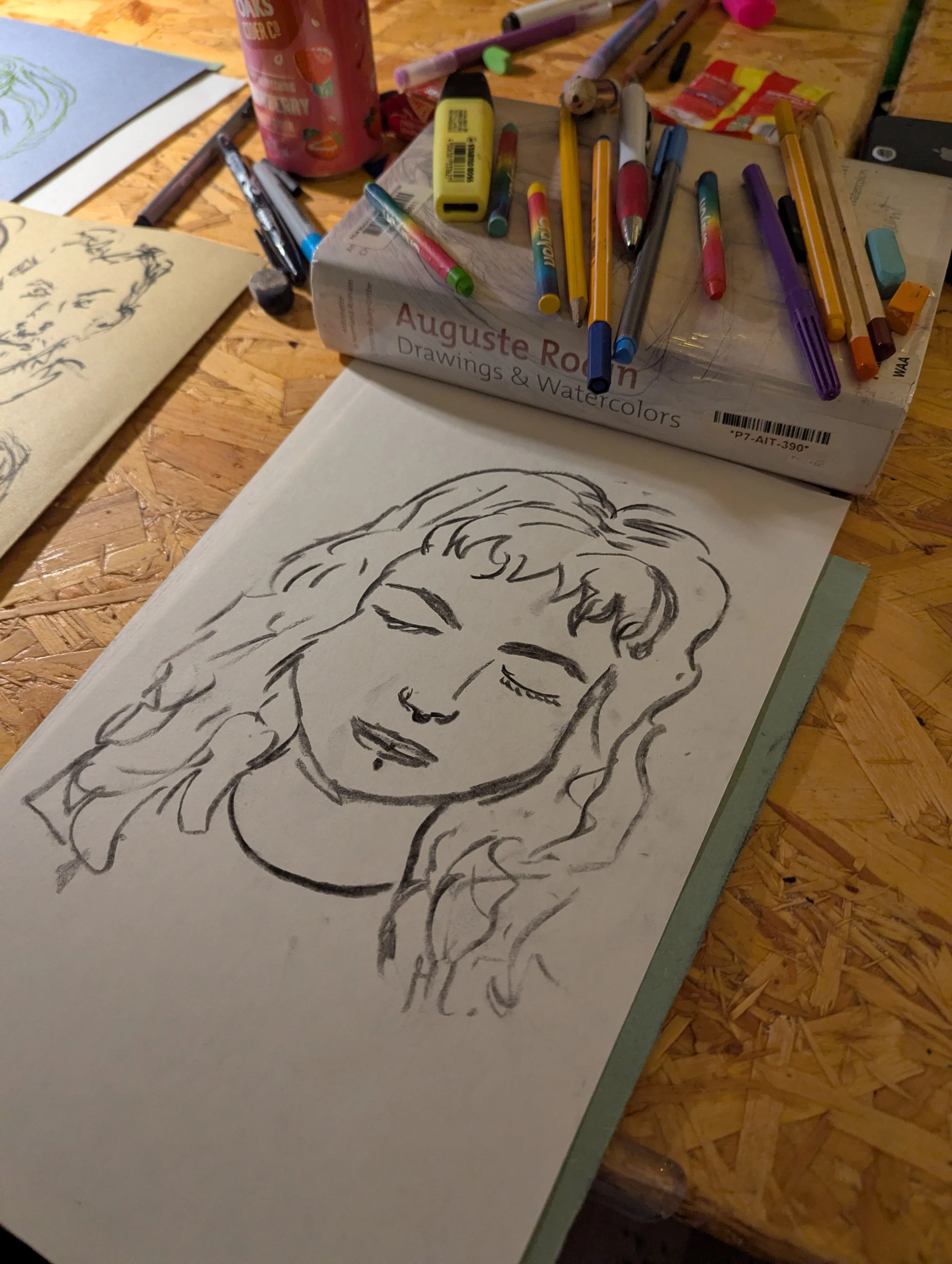 Line drawing of a woman's face with closed eyes and wavy hair, on a sheet of paper on a wooden table surrounded by art supplies and drawing tools.