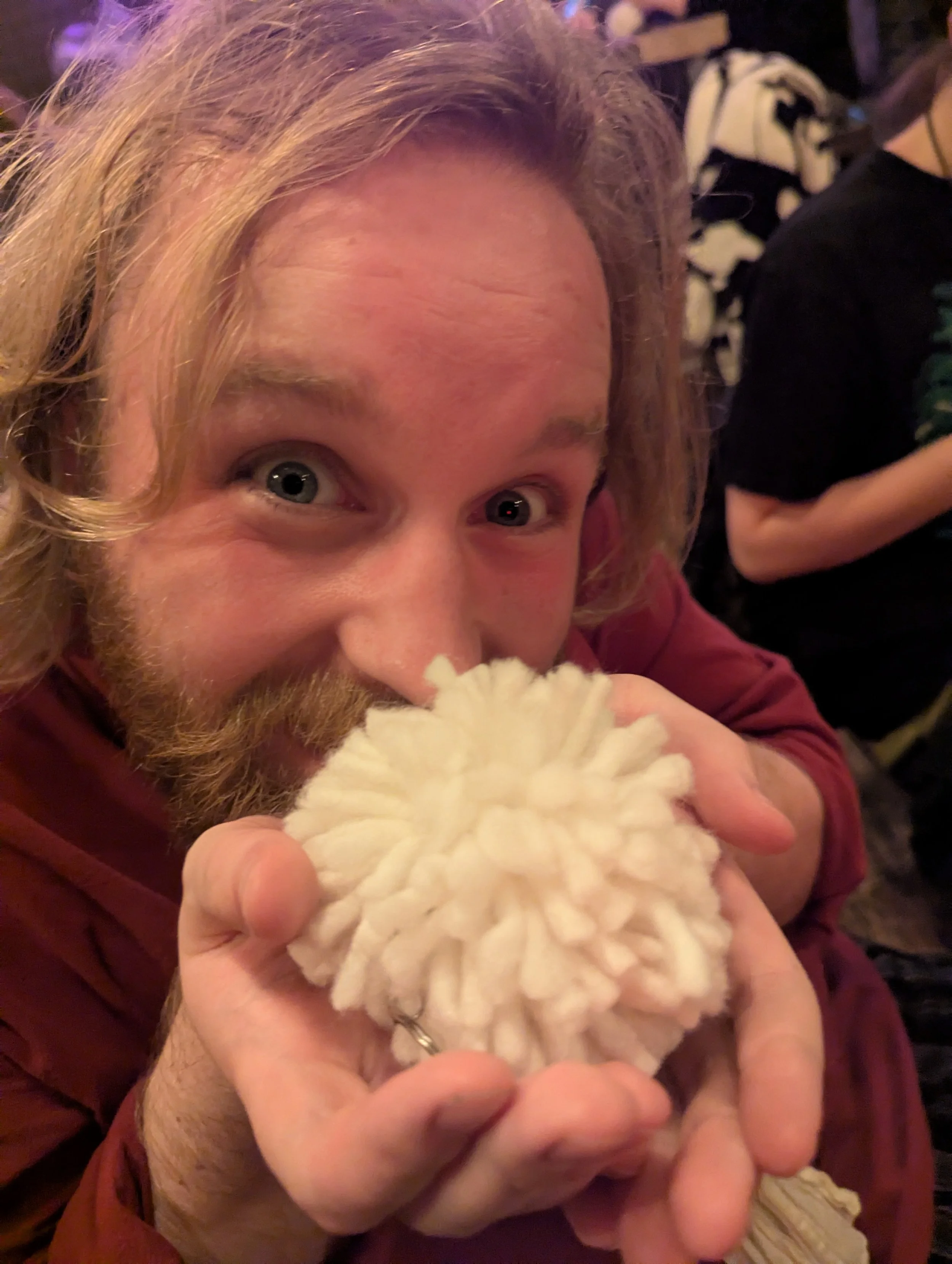 A man with long blond hair and a beard smiling and holding a large white fluffy cotton ball close to his face.