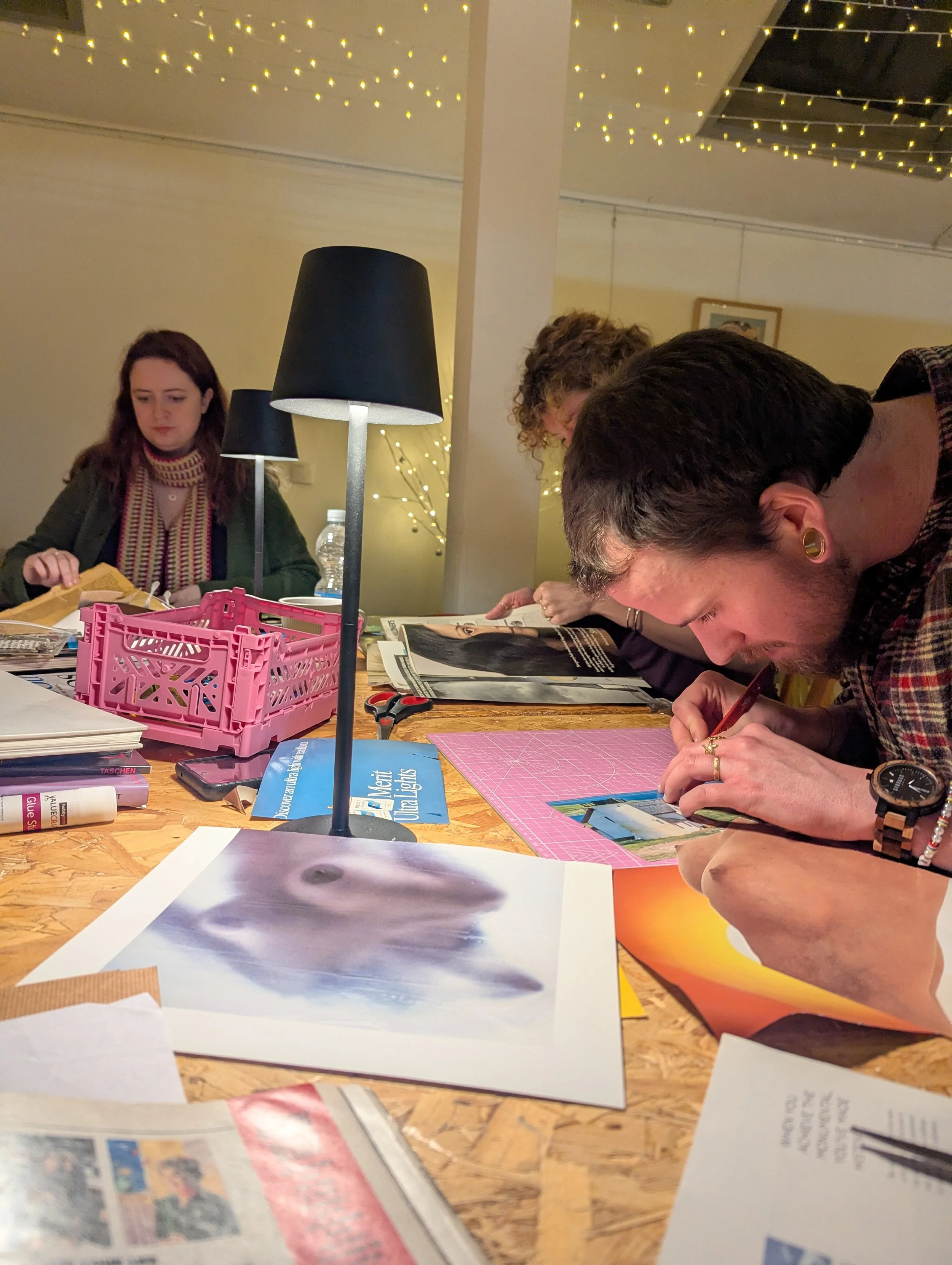 People working on arts and crafts at a wooden table decorated with magazines, paper, scissors, and a pink storage basket. Dim lighting with a black lampshade and string lights in the background.