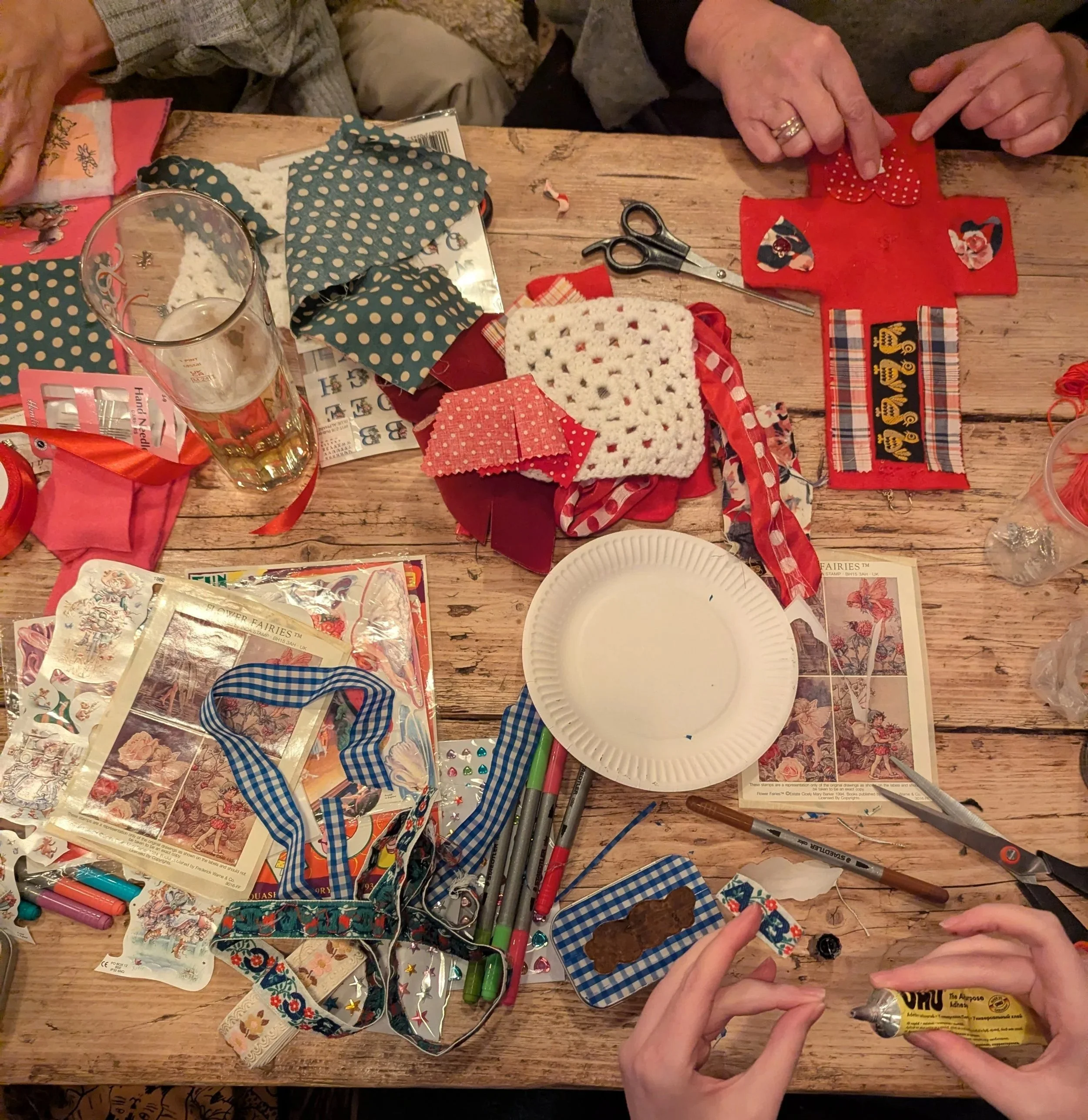 A wooden table cluttered with craft supplies, fabric pieces, scissors, markers, design sheets, a paper plate, and a person's hands working with a tube of glue, surrounded by people working on a craft project.