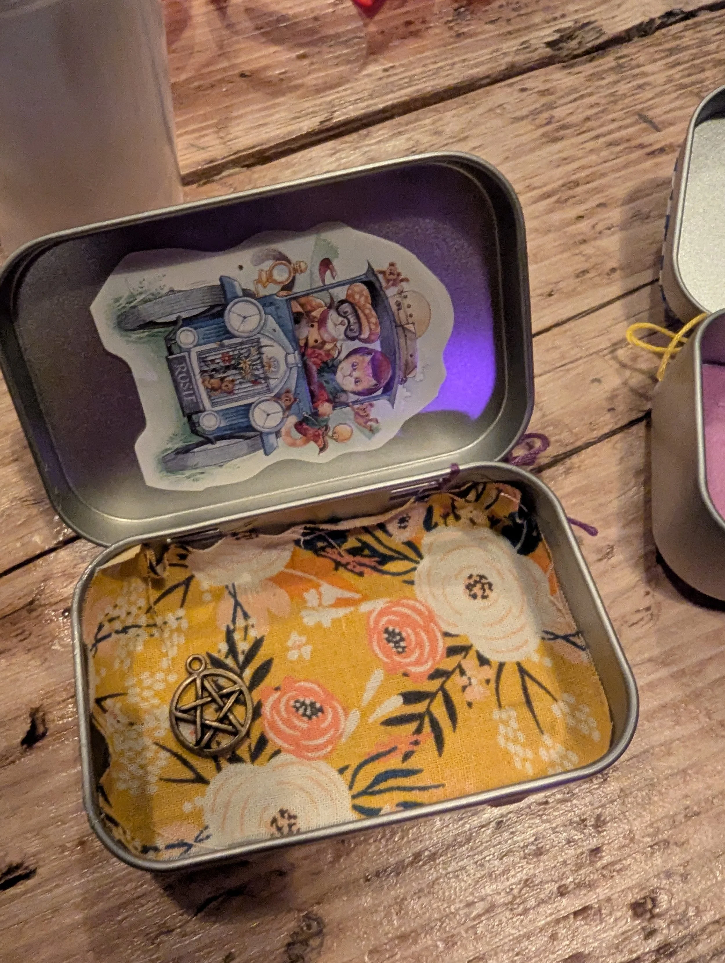 Open metal tin box with a decorative illustration inside, a yellow floral patterned fabric lining, and a small metallic pentacle charm. Other metal tins are partially visible nearby on a wooden surface.