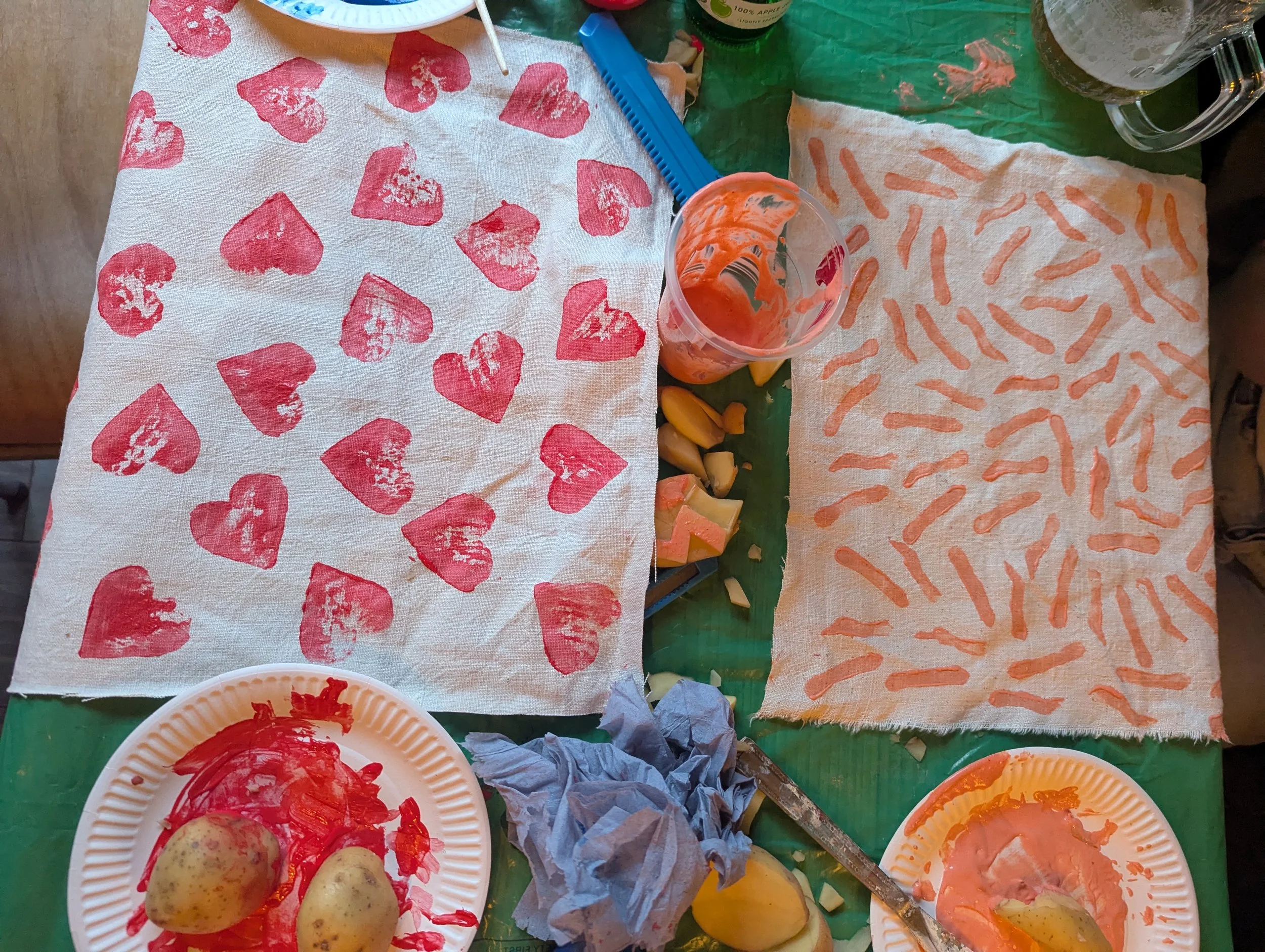 Party table with red heart and orange striped paper towels, a glass of orange juice, peeled potatoes, and cheese and melon pieces.