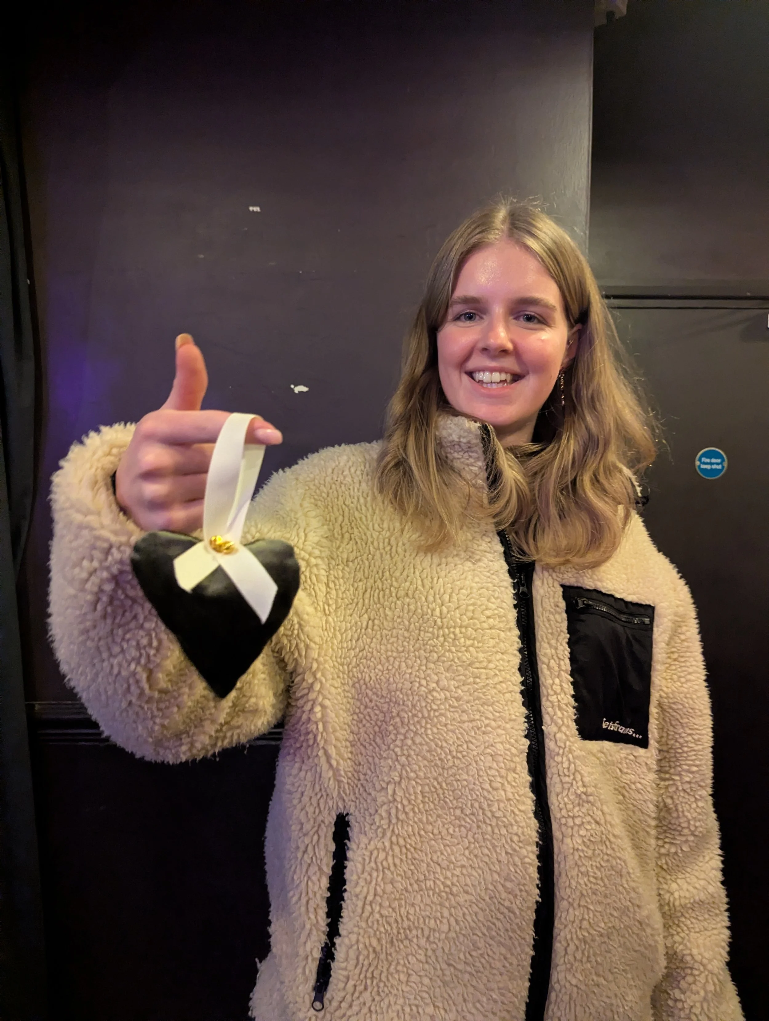 A smiling woman in a beige fleece coat holding a small black heart-shaped pillow with a white ribbon and gold decoration, giving a thumbs-up gesture.