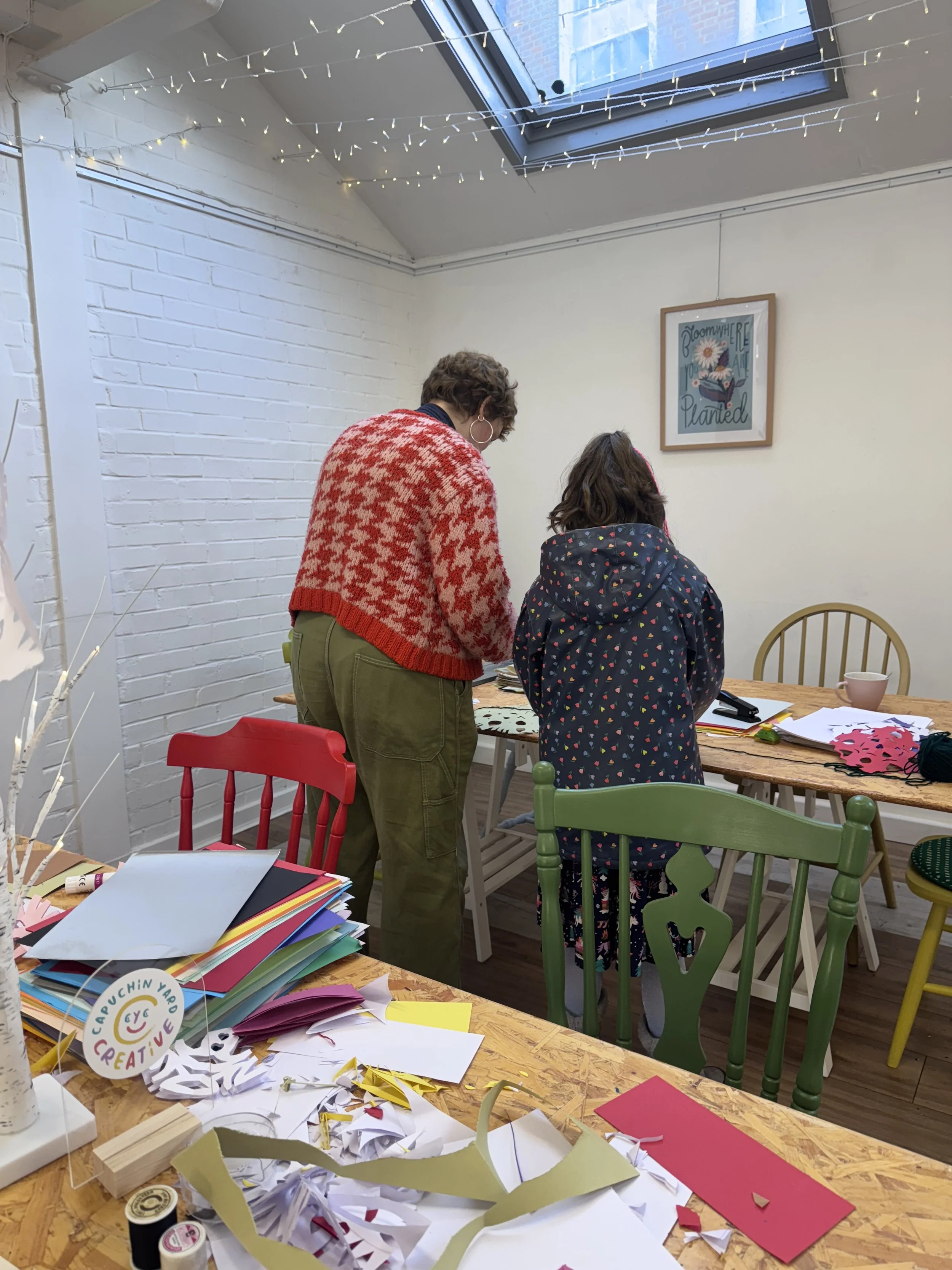 Two women working on arts and crafts at a table in a creative space decorated with fairy lights and colorful chairs, with craft materials scattered on the table.